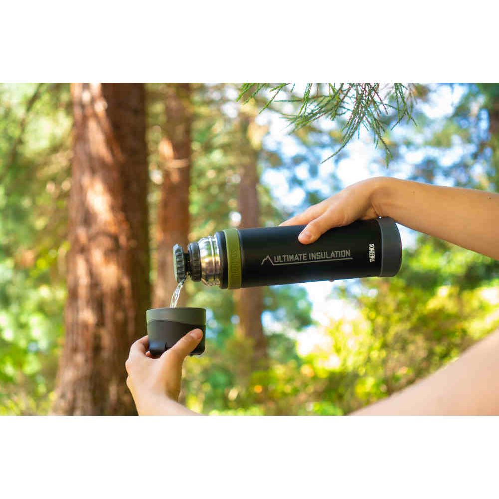 Eine Person gießt Wasser aus einer schwarzen Thermoskanne in eine Tasse, umgeben von einem sonnigen Wald.