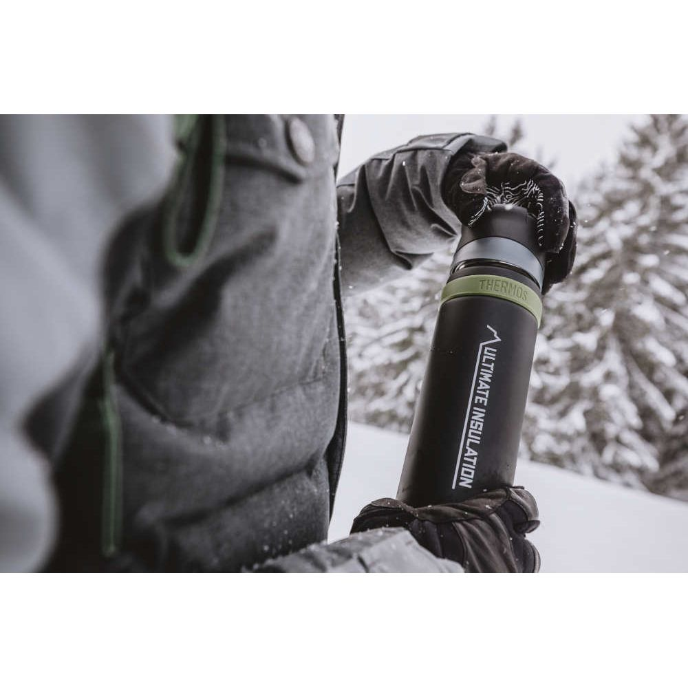 Person in Winterkleidung hält eine schwarze Thermoskanne im verschneiten Wald.