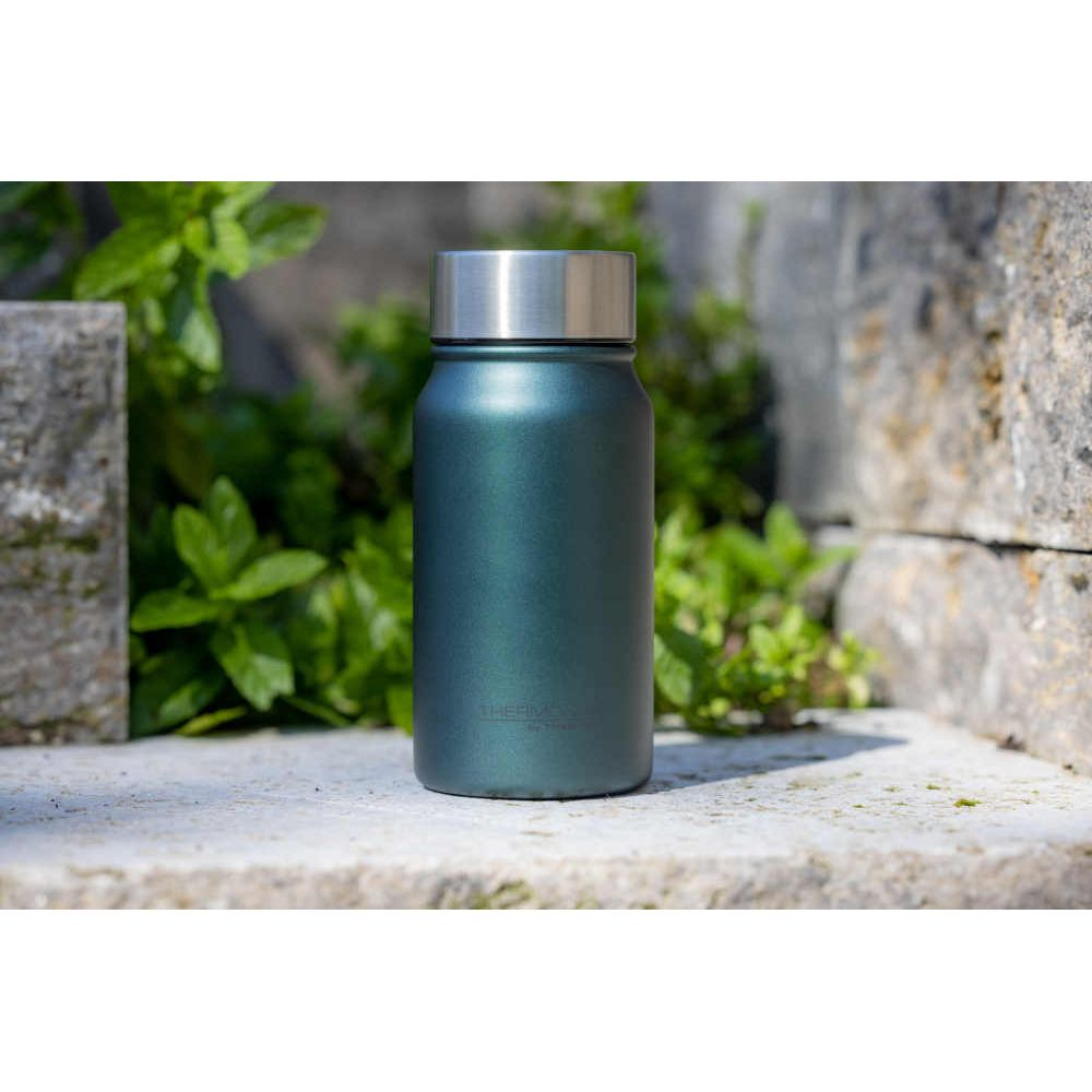 Eine metallische, dunkelgrüne Thermosflasche mit silbrigem Deckel steht auf einer Steinplatte im Freien vor grünen Blättern.