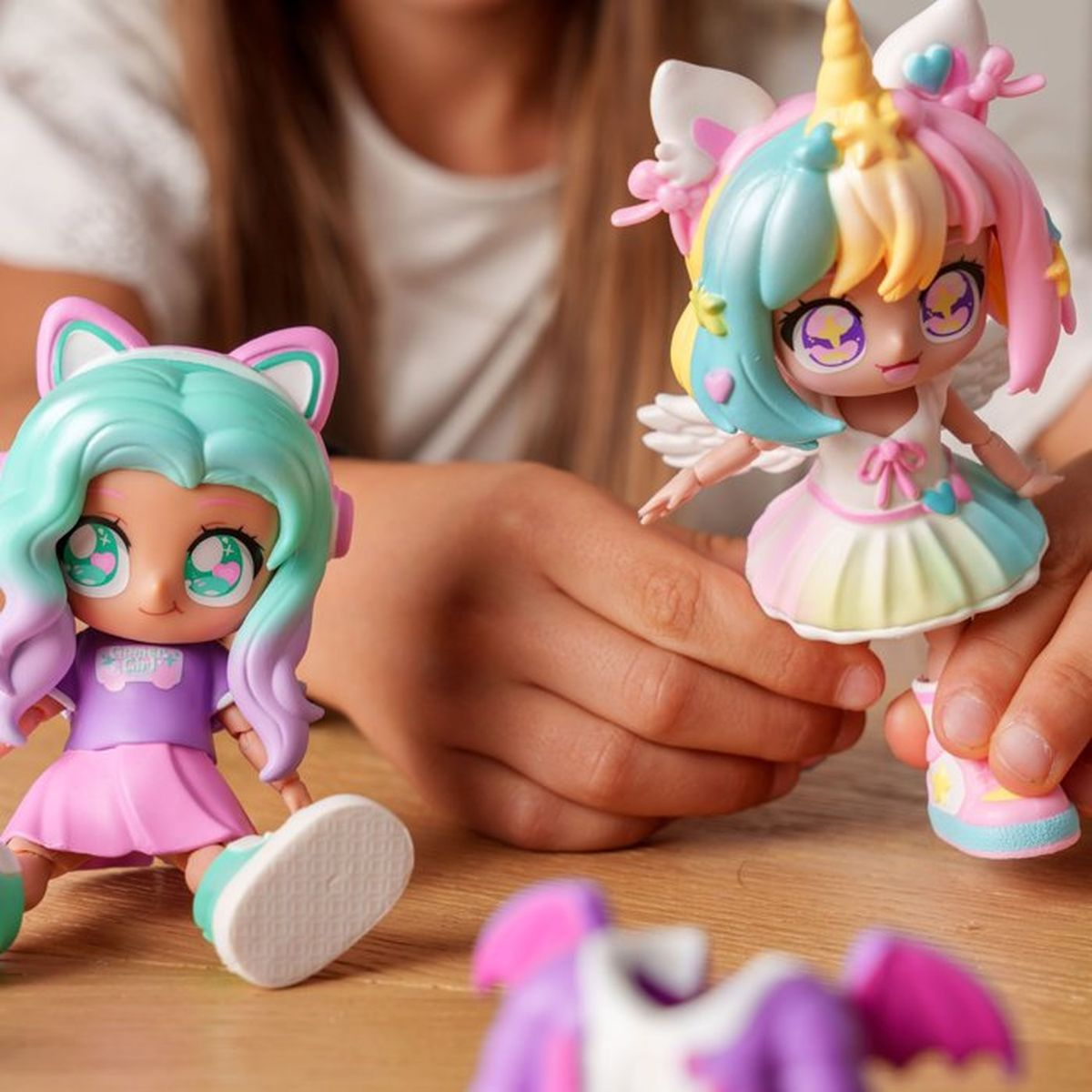 Ein Kind hält zwei bunte, anime-artige Spielzeugpuppen mit großen Augen auf einem Holztisch.