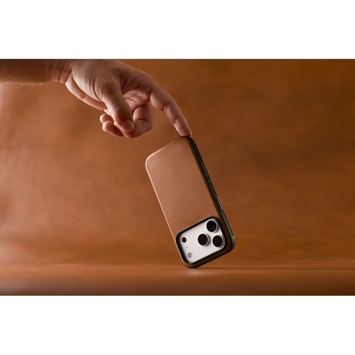Eine Hand berührt leicht eine braune Lederhülle auf einem Smartphone vor warmem braunen Hintergrund.