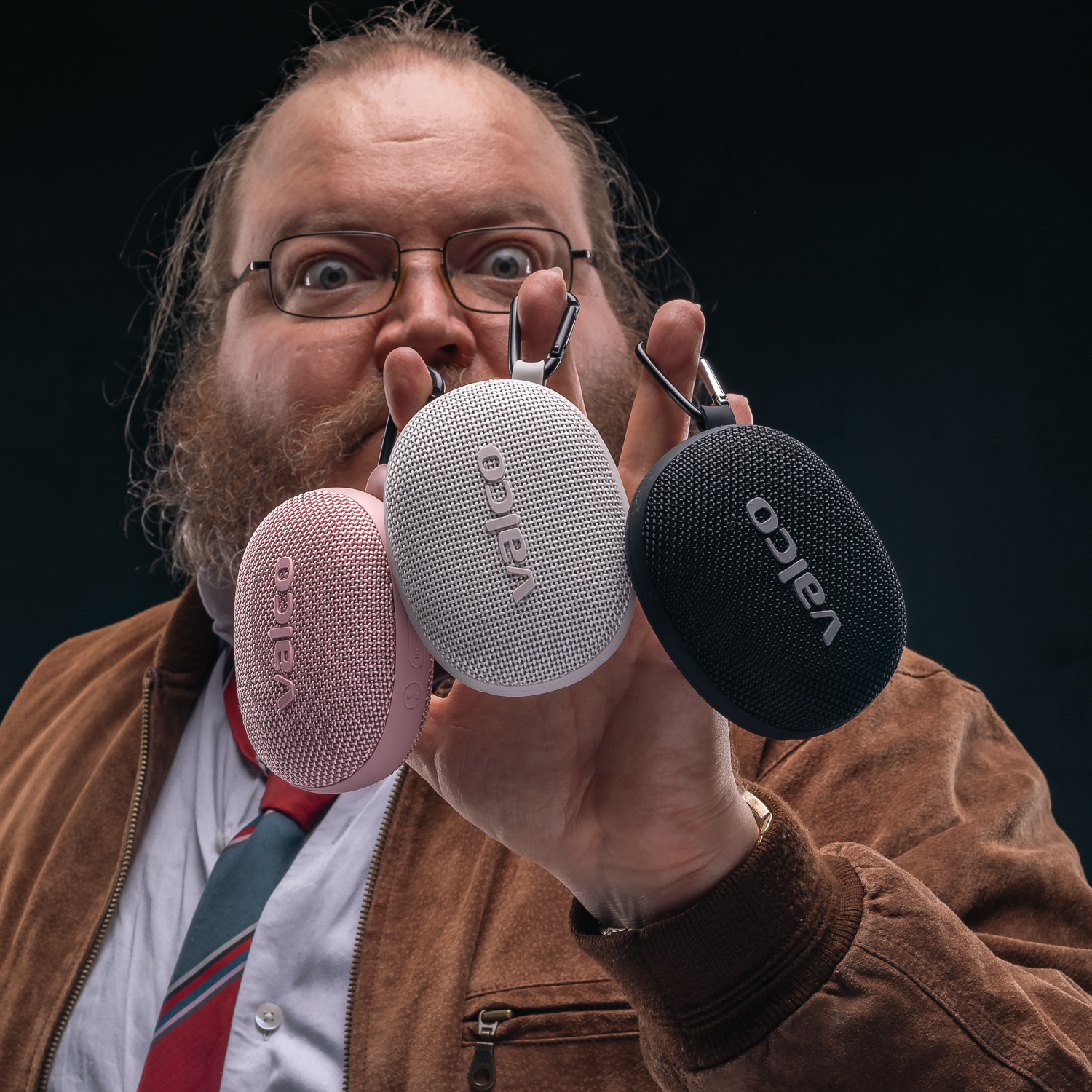 Ein Mann mit Brille und Bart hält drei ovale Valco-Lautsprecher in Rosa, Weiß und Schwarz in die Kamera.