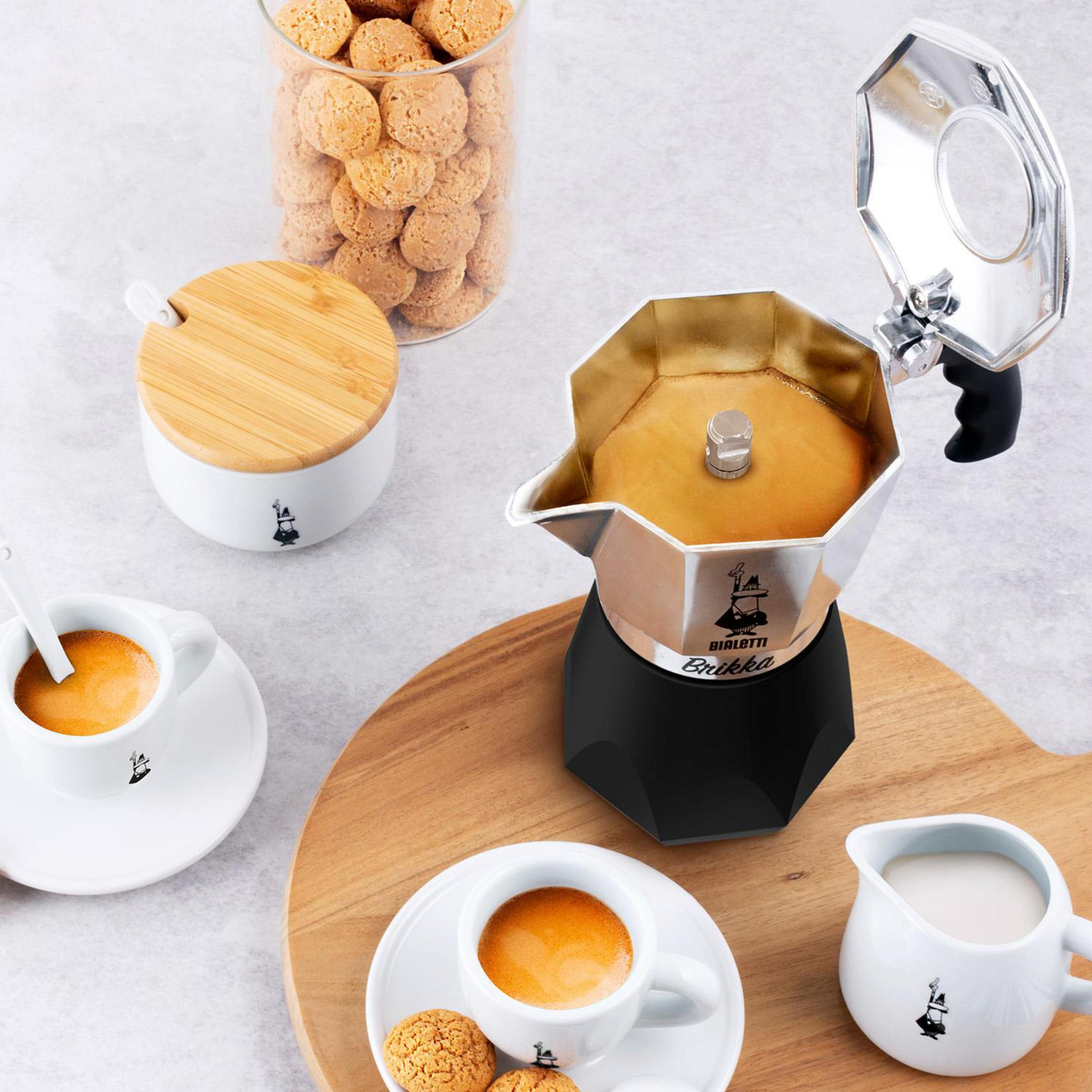 Otwarta kawiarka, dwie filiżanki espresso, dzbanek na mleko, cukiernica i ciasteczka na drewnianej desce.