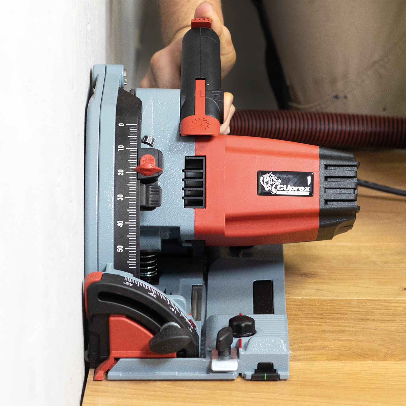 Hand hält eine rot-graue Tauchsäge nahe an einer weißen Wand auf einem Holzboden.