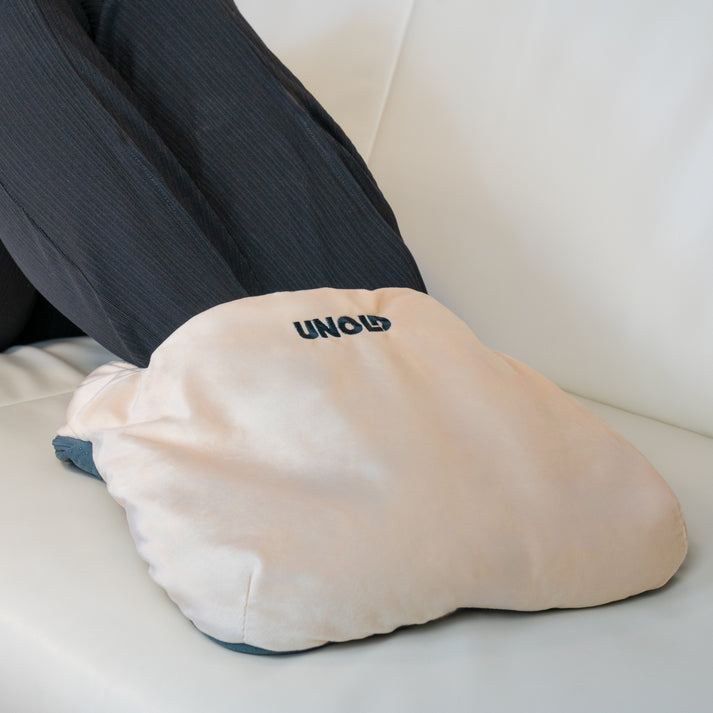 Ein beige Unold Fußwärmer liegt auf einer weißen Couch mit dunklen Hosen darin.