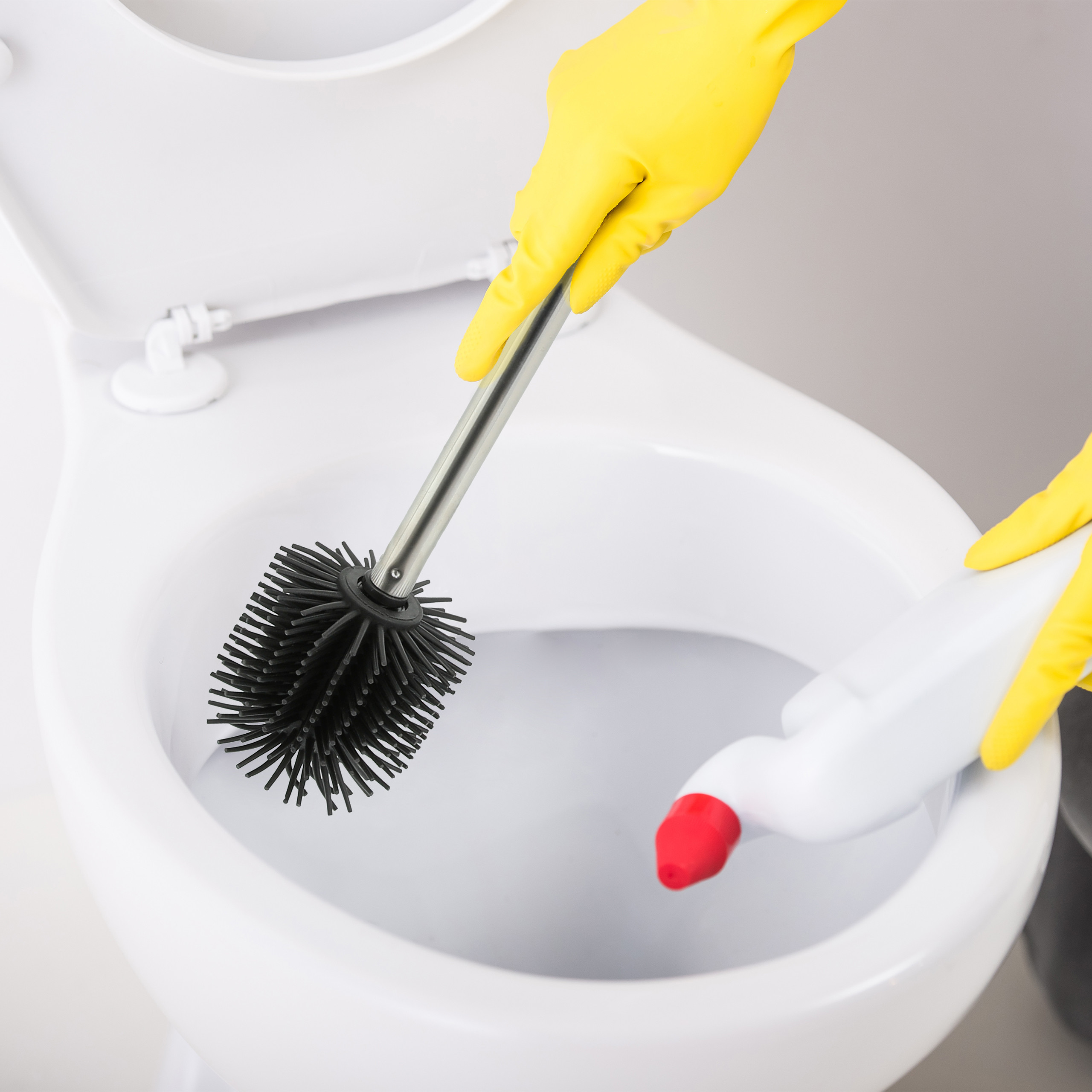Gelbe Handschuhe reinigen eine weiße Toilette mit einer schwarzen Bürste und einer Flasche Reiniger.