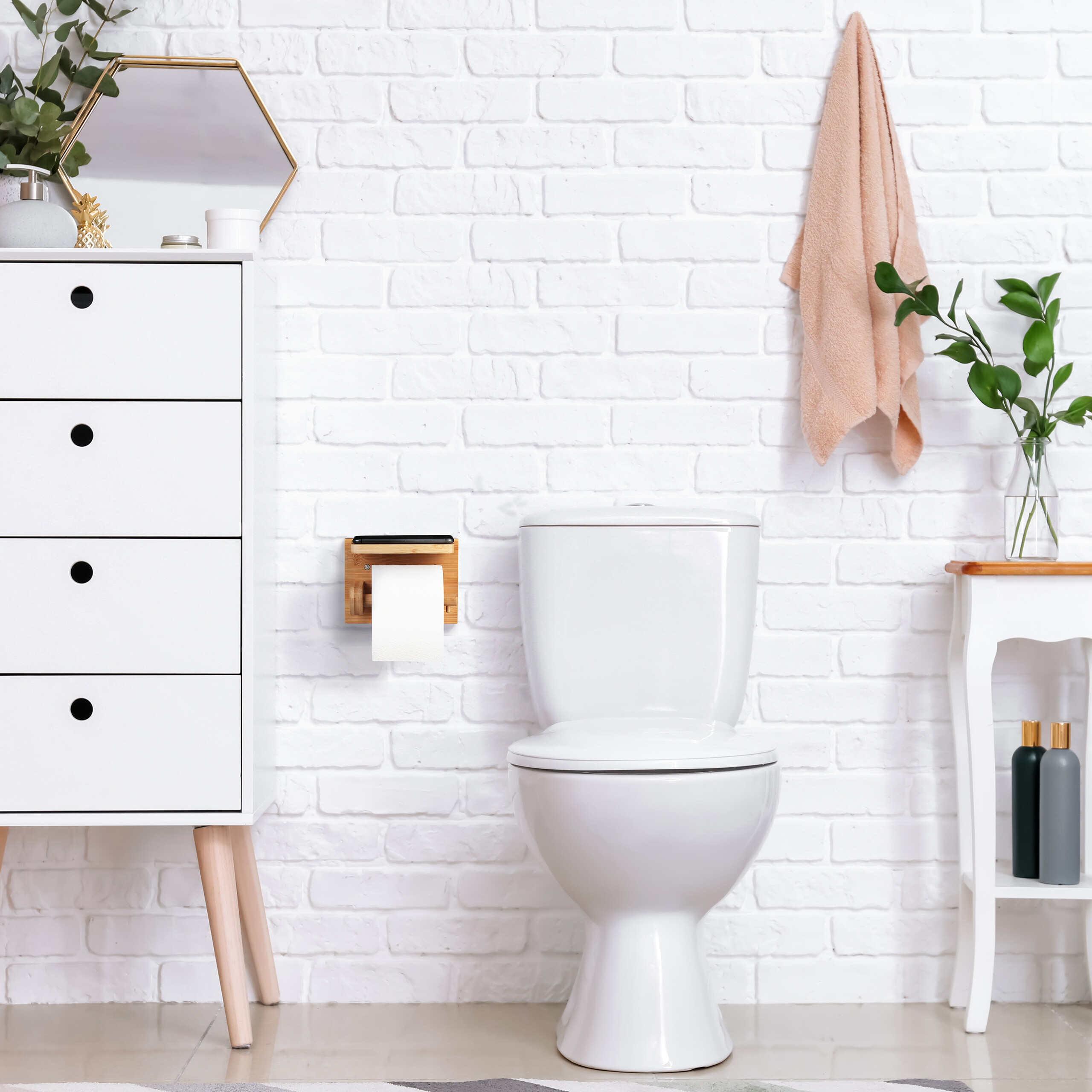 Eine weiße Toilette steht vor einer weißen Backsteinwand. Links ein Schrank, rechts ein Tisch mit Pflanzen und Handtuch.