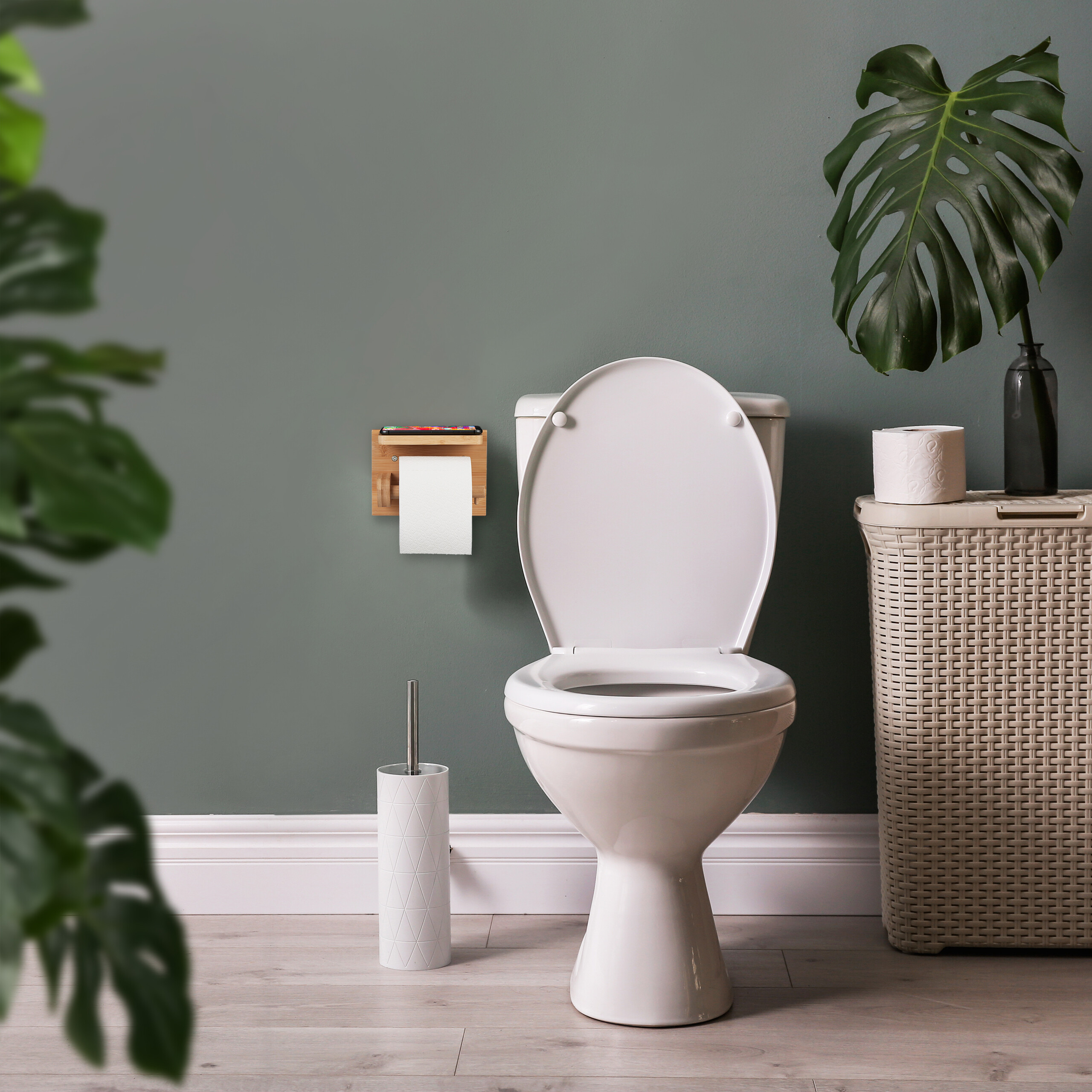 Weißes WC mit offenem Deckel an grüner Wand, Holzhalter, weißer Bürstenhalter und Korb mit Pflanze.