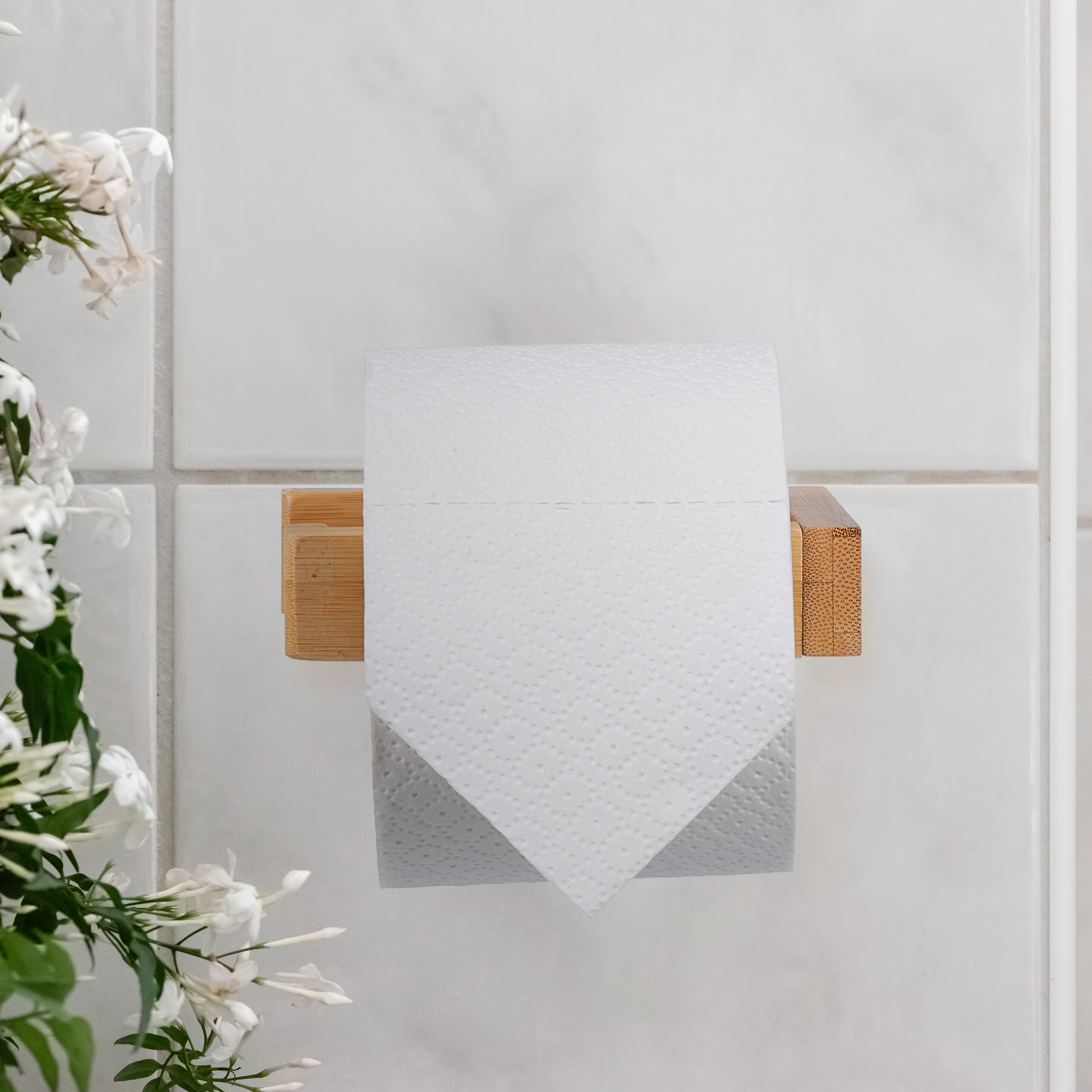 Eine Toilettenpapierrolle hängt an einem Holzhalter an weißen Fliesen, links sind weiße Blüten zu sehen.