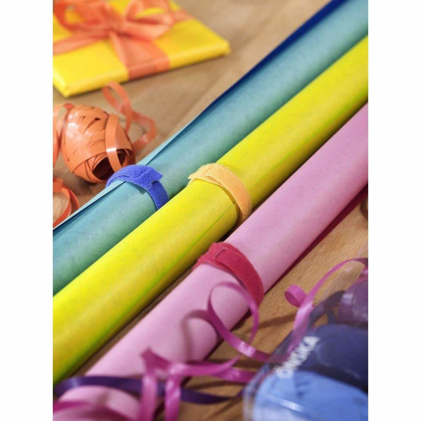 Gerollte Geschenkpapierbögen in Blau, Gelb und Rosa mit farbigen Bändern auf Holz.