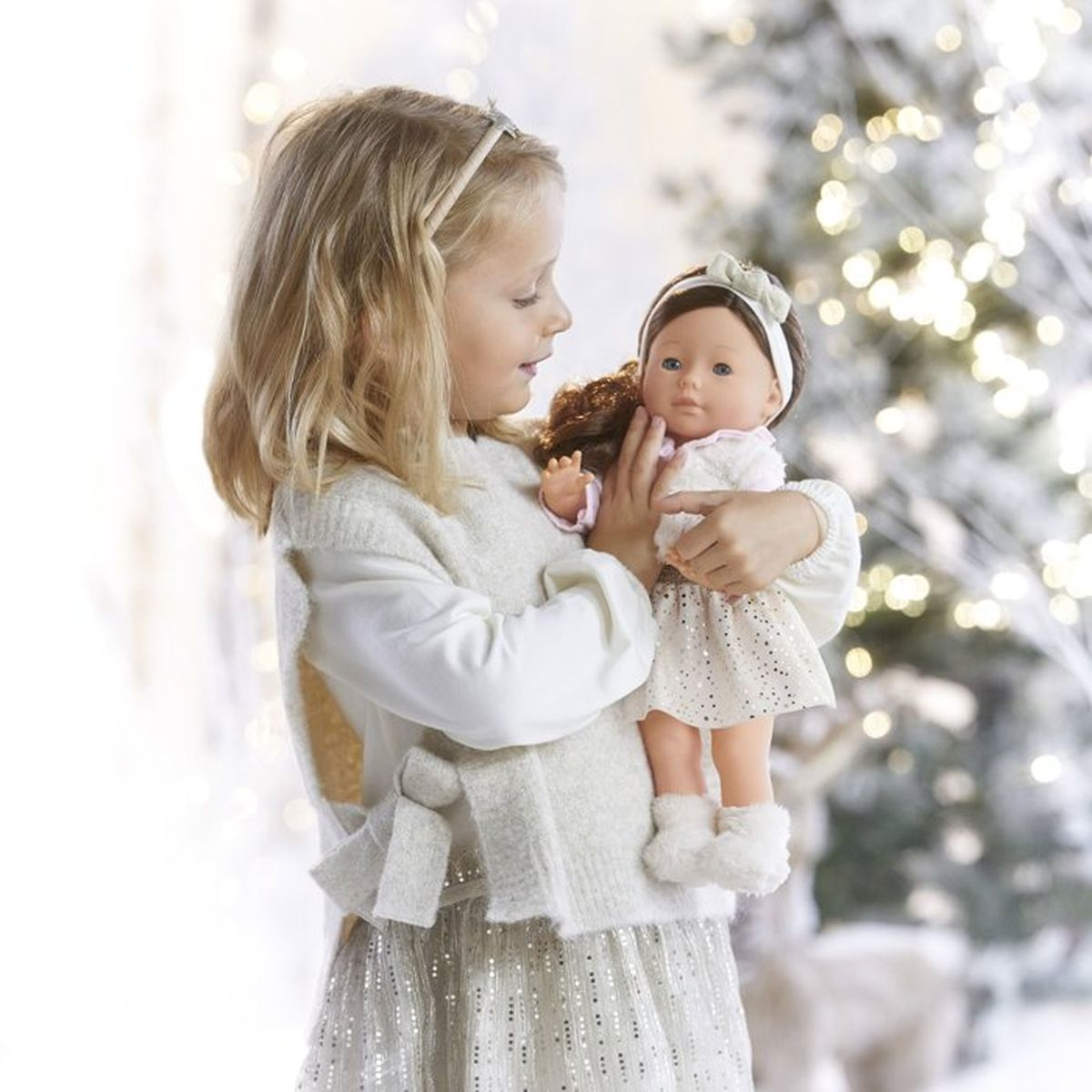 Ein blondes Mädchen im weißen Strickpullover hält eine Puppe vor einem verschneiten Weihnachtsbaum mit Lichtern.