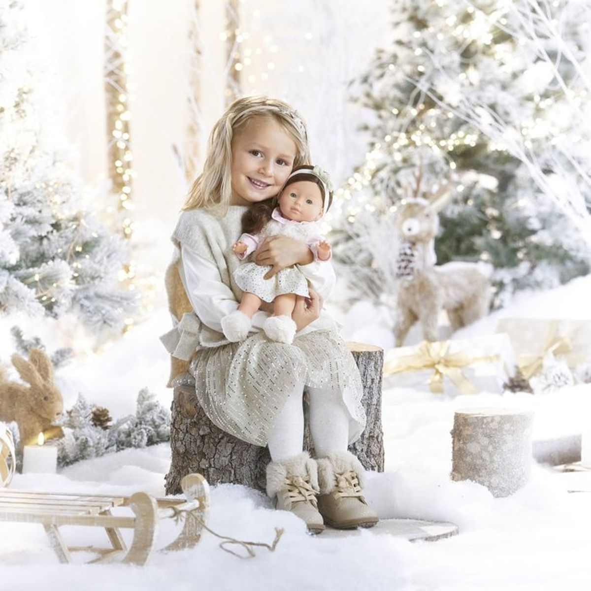 Ein blondes Mädchen sitzt auf einem Baumstumpf in einer verschneiten Winterlandschaft und hält eine Puppe im Arm.