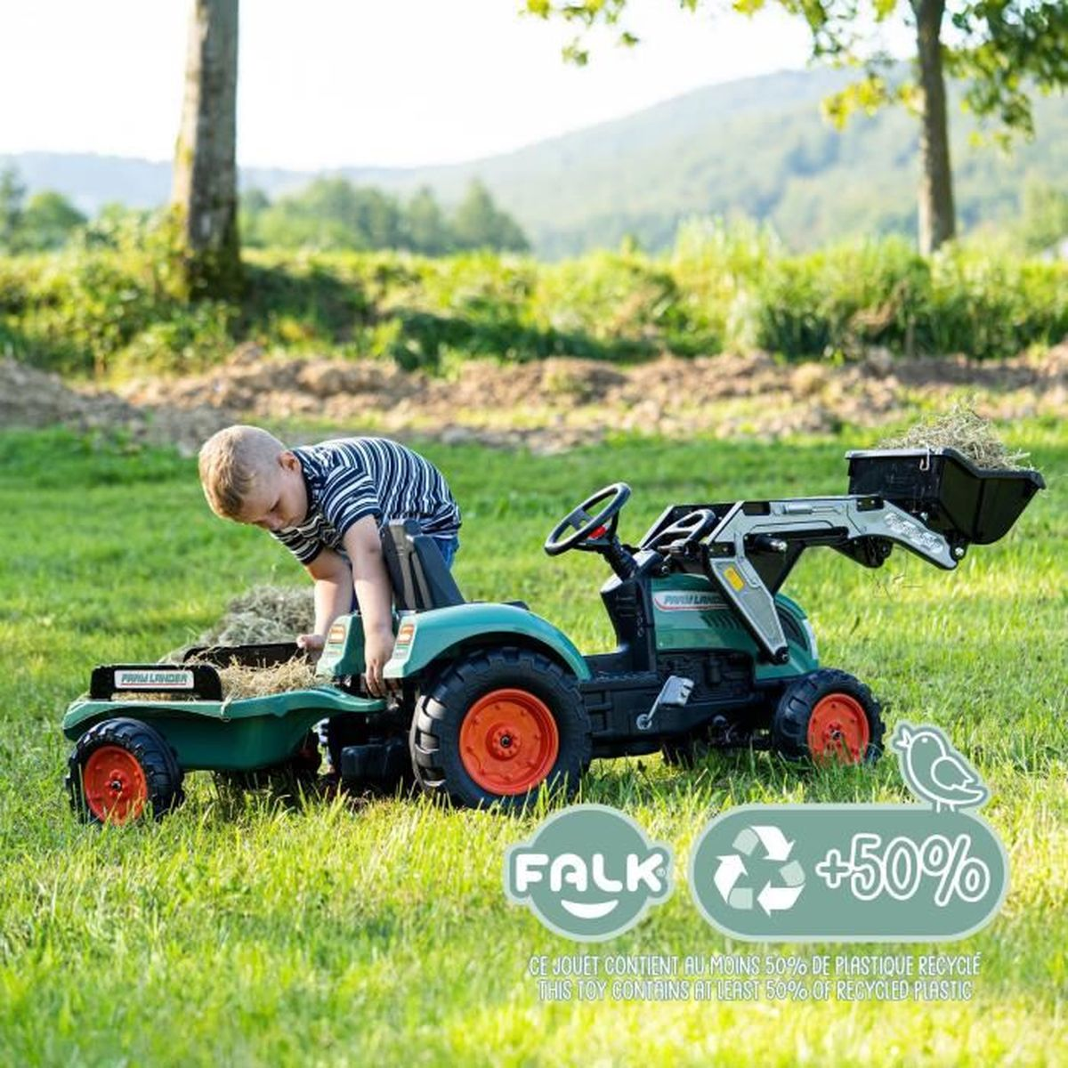 Ein kleiner Junge spielt mit einem grünen Spielzeugtraktor und Anhänger auf einer Wiese.