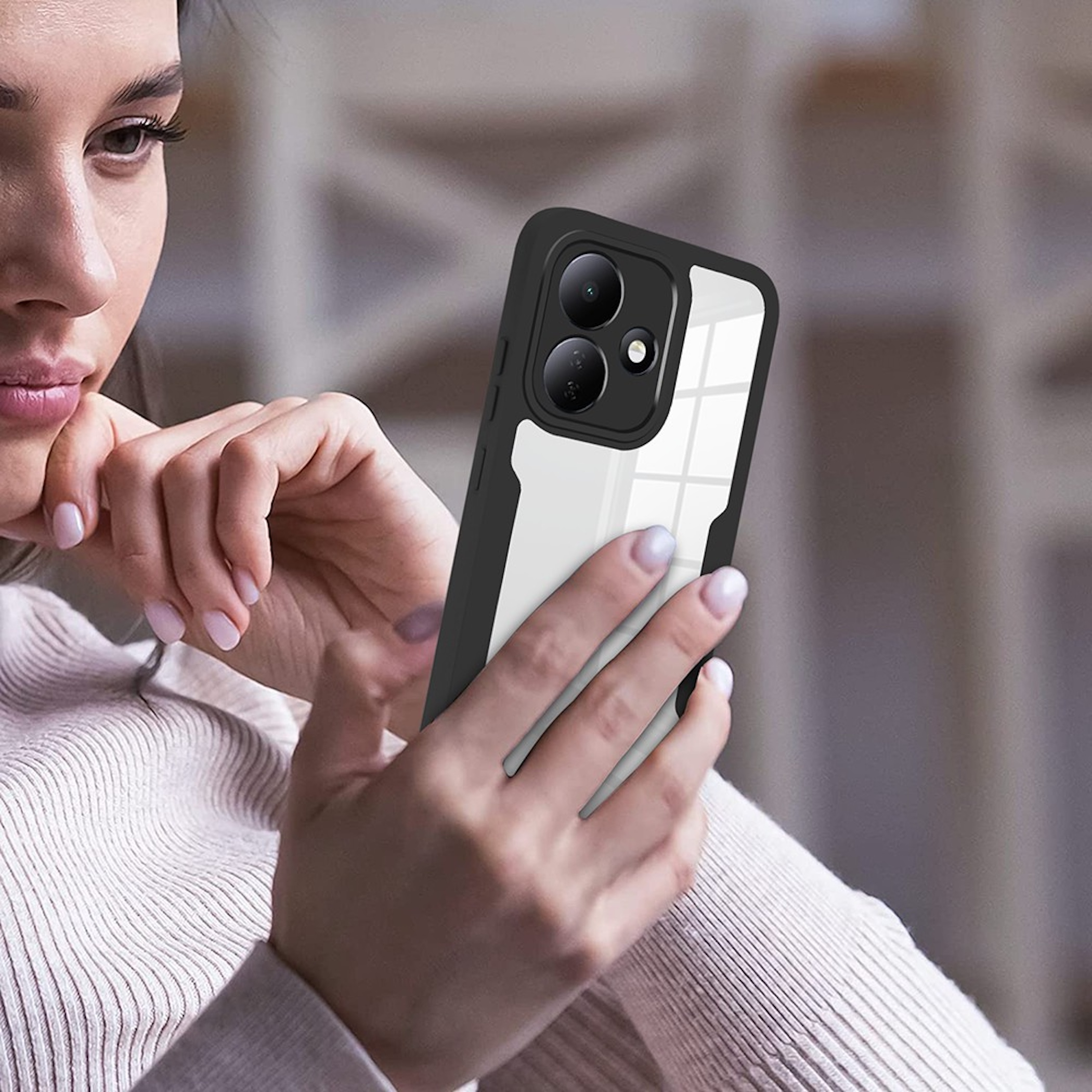 Eine Frau hält ein Smartphone mit schwarzem Bumper und klarer Rückseite, sitzend in einem Raum mit verschwommenem Hintergrund.