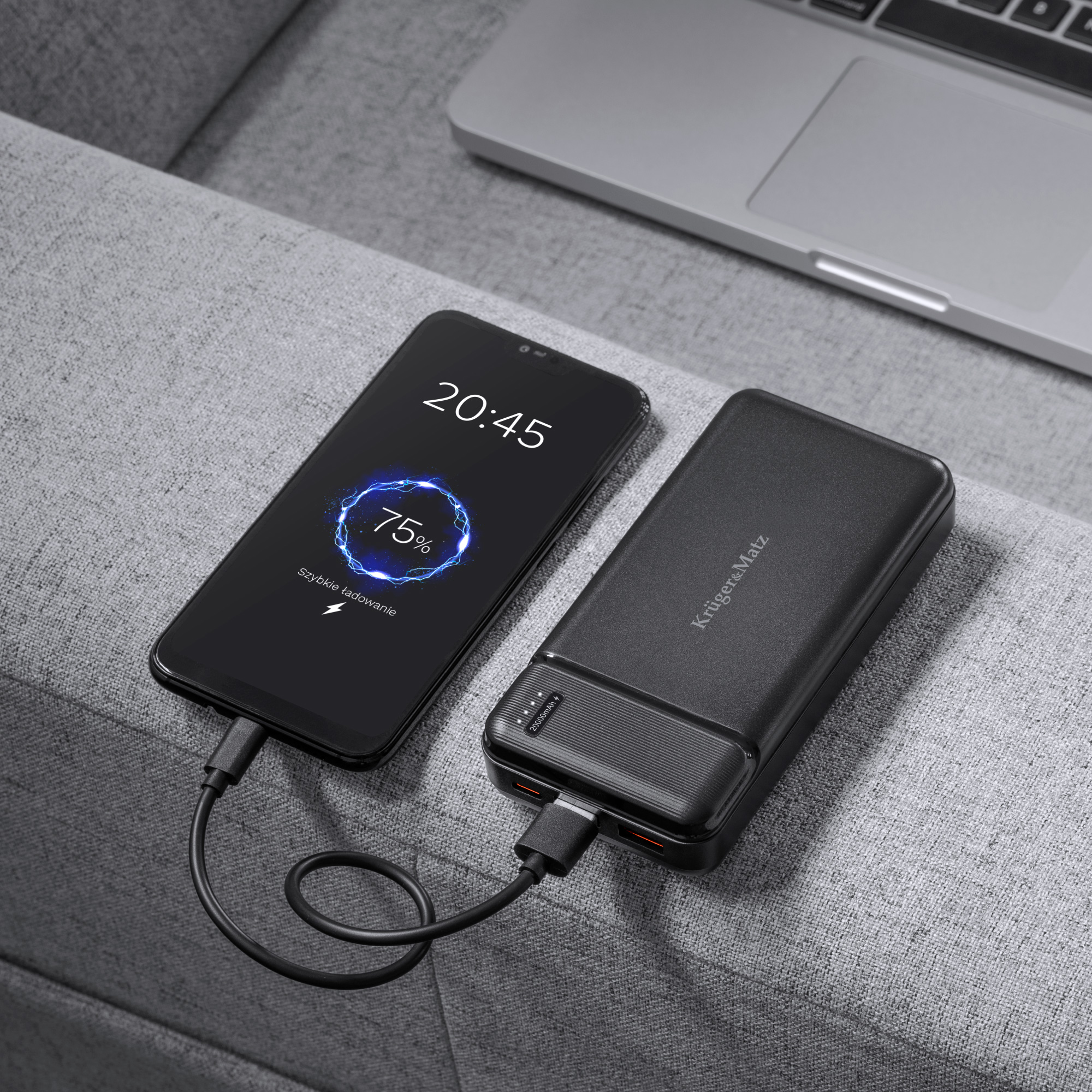 Smartfon ładuje się z czarnego powerbanka na szarej kanapie, w tle widać laptopa.
