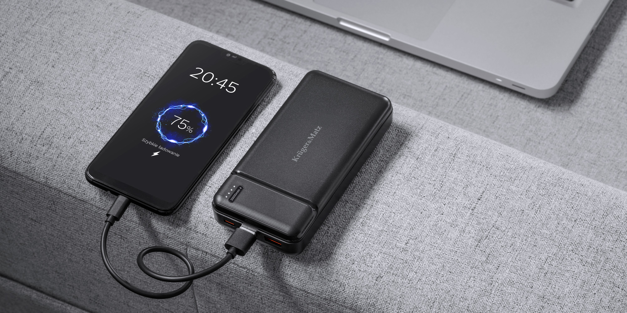 Smartfon jest ładowany przez czarny powerbank na szarej kanapie, w tle widać laptopa.