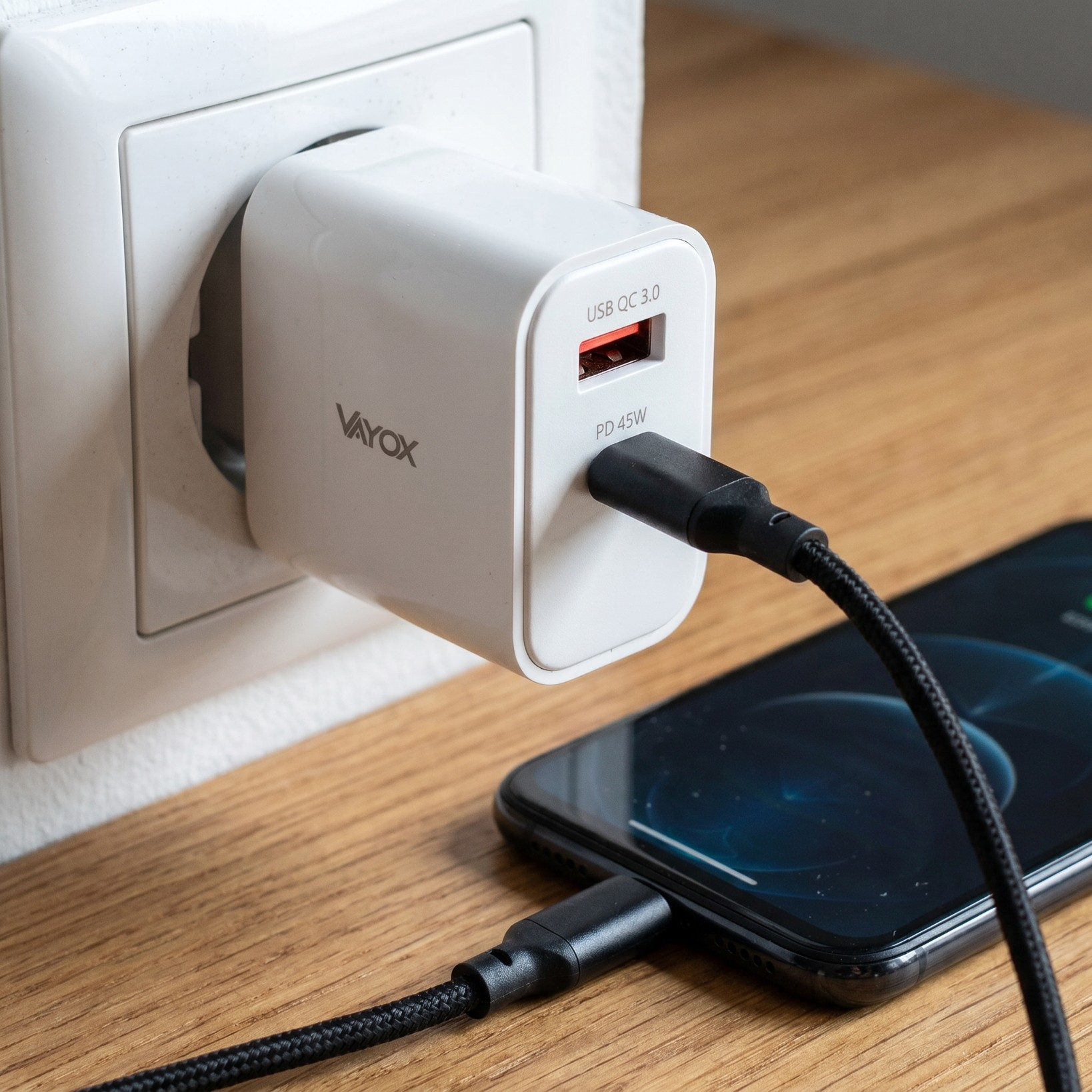 Fehér Vayox USB töltő fali aljzatba dugva, fekete kábellel okostelefont tölt egy fa asztalon.