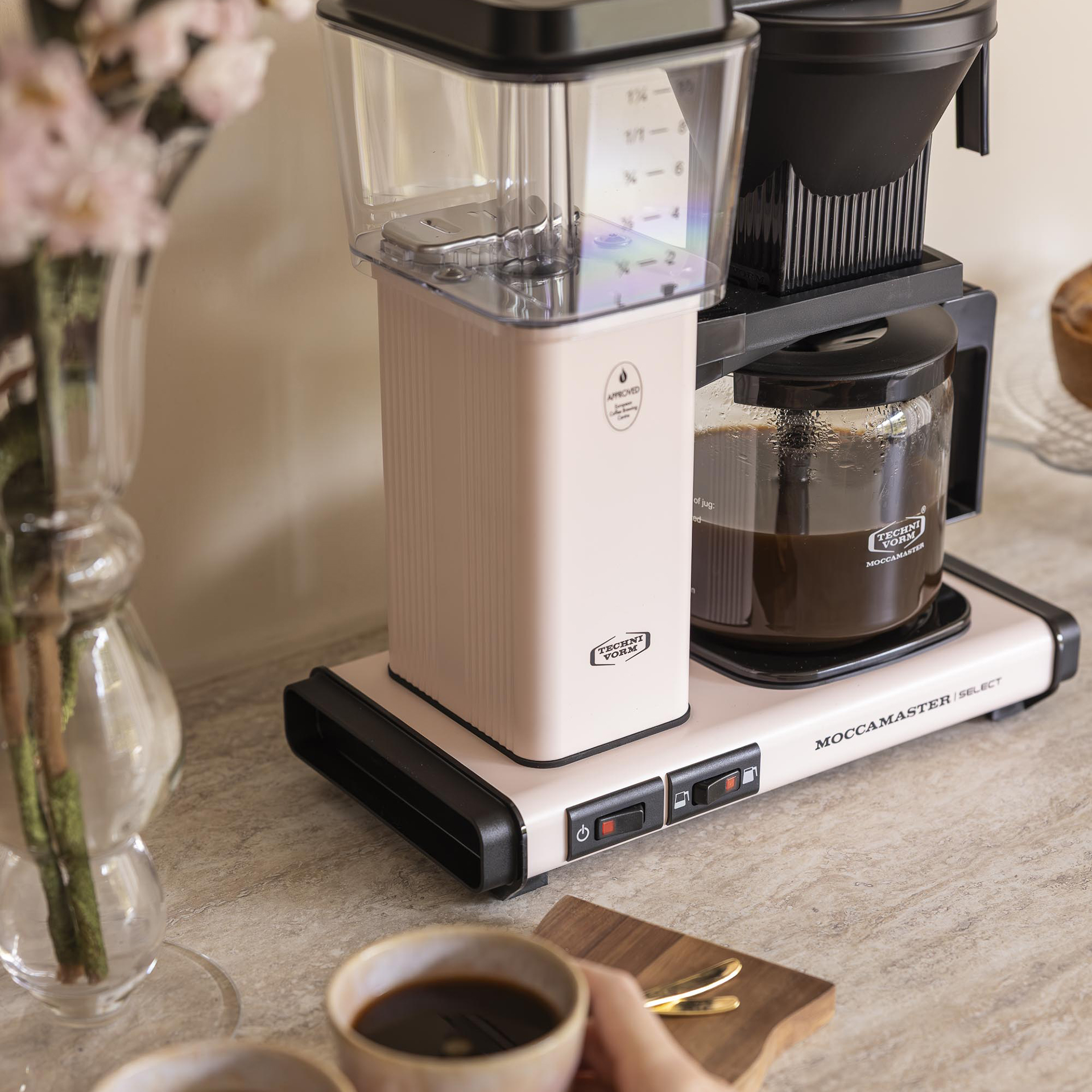 Una macchina da caffè Moccamaster rosa si trova su un piano di cucina accanto a un vaso di fiori e a una tazza di caffè.