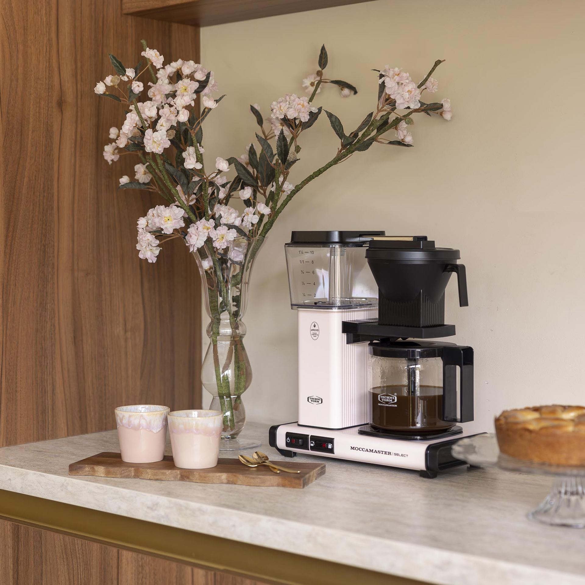 Una macchina da caffè rosa, due tazze e un vaso con fiori rosa sono su un bancone da cucina.