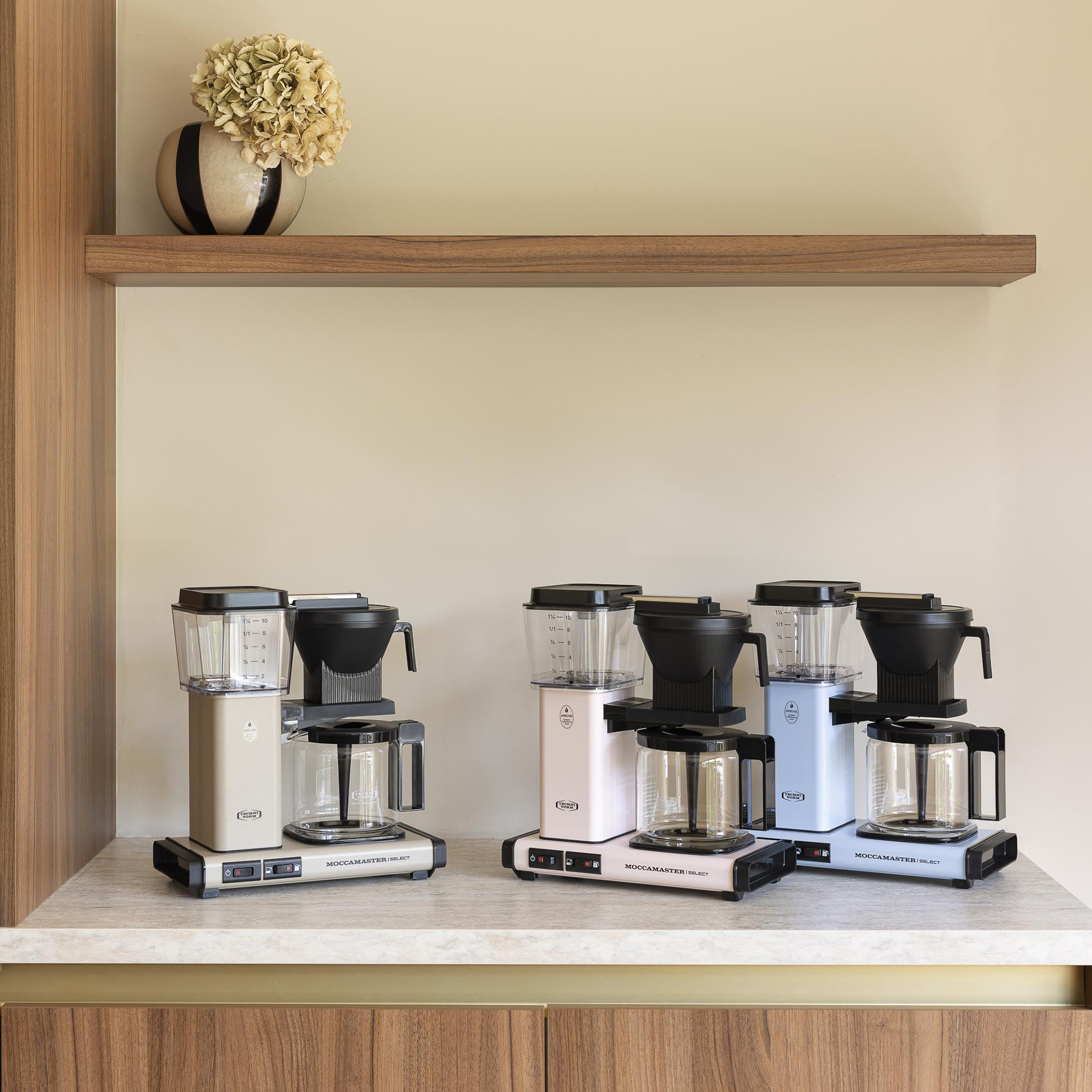 Tre macchine da caffè Technivorm Moccamaster beige, rosa e blu sono su un bancone sotto un ripiano in legno.