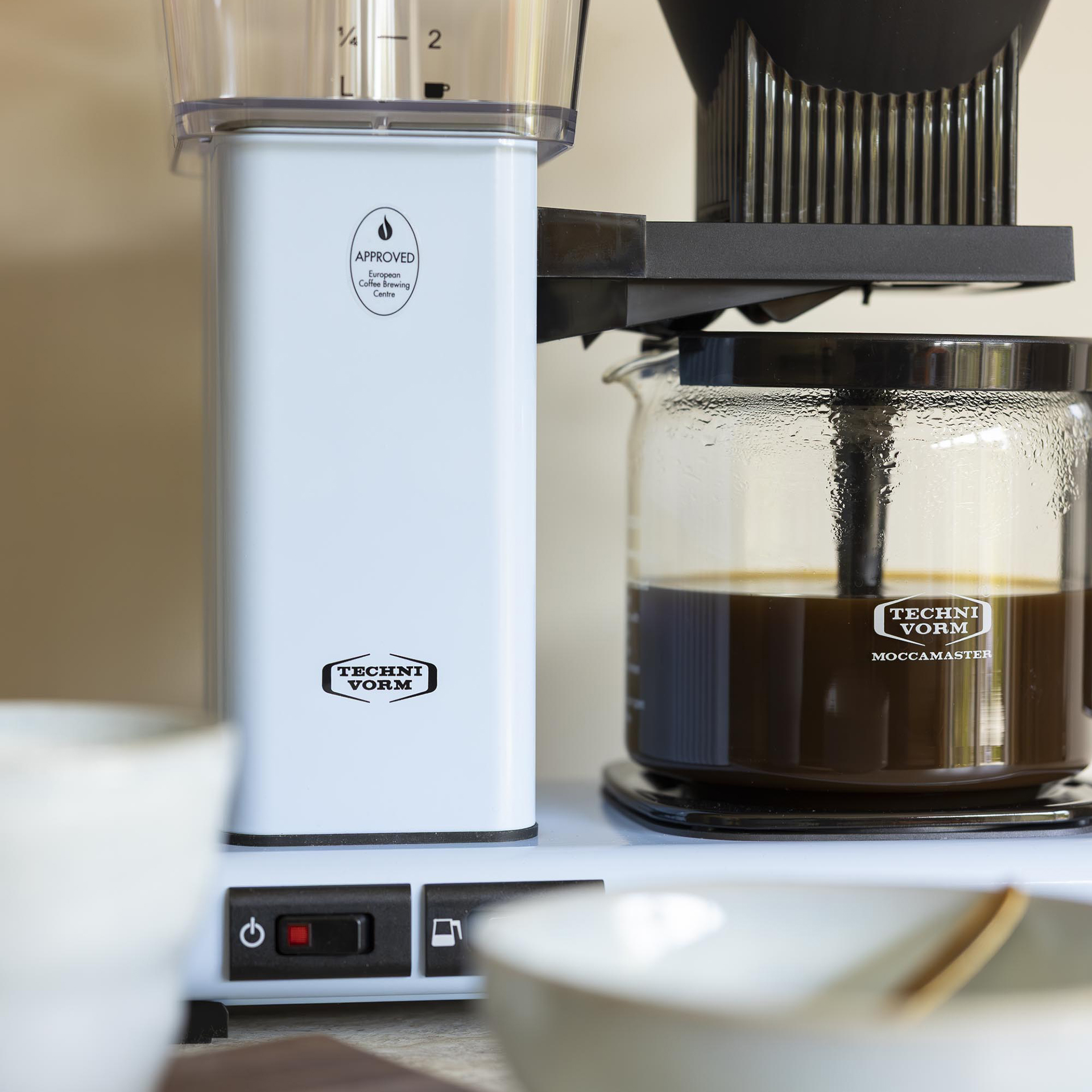 Une cafetière Technivorm Moccamaster bleu clair prépare du café, avec des tasses au premier plan.