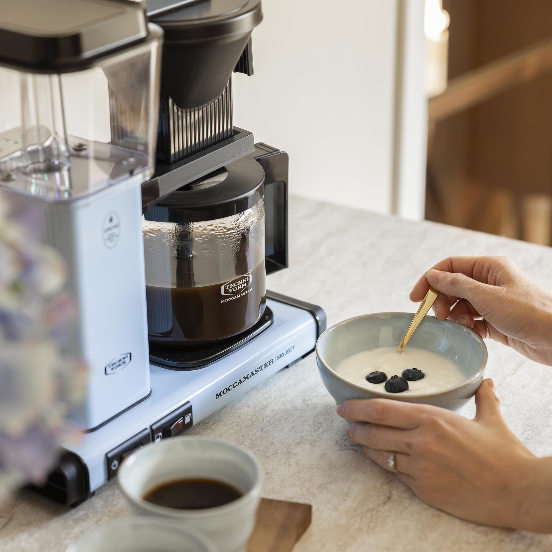 Une cafetière bleu clair sur le comptoir pendant qu'une personne mange du yaourt aux myrtilles.