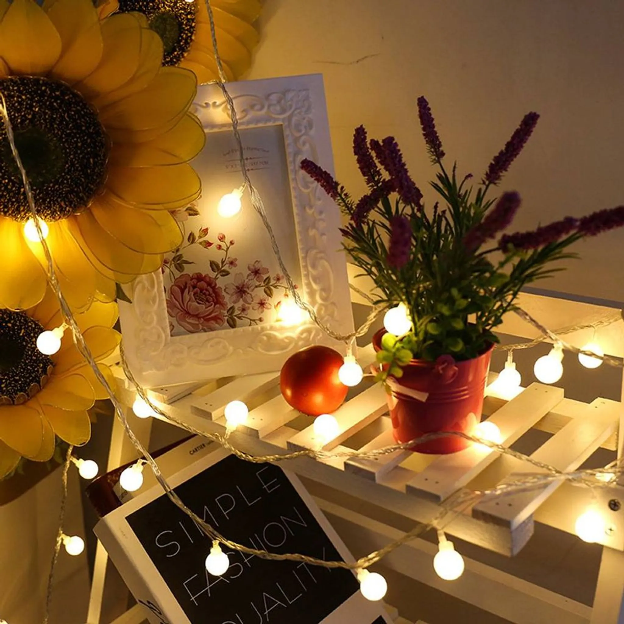 Ein Holzregal mit Lichterketten, einem Bilderrahmen, einer Topfpflanze, einer Tomate und Büchern.