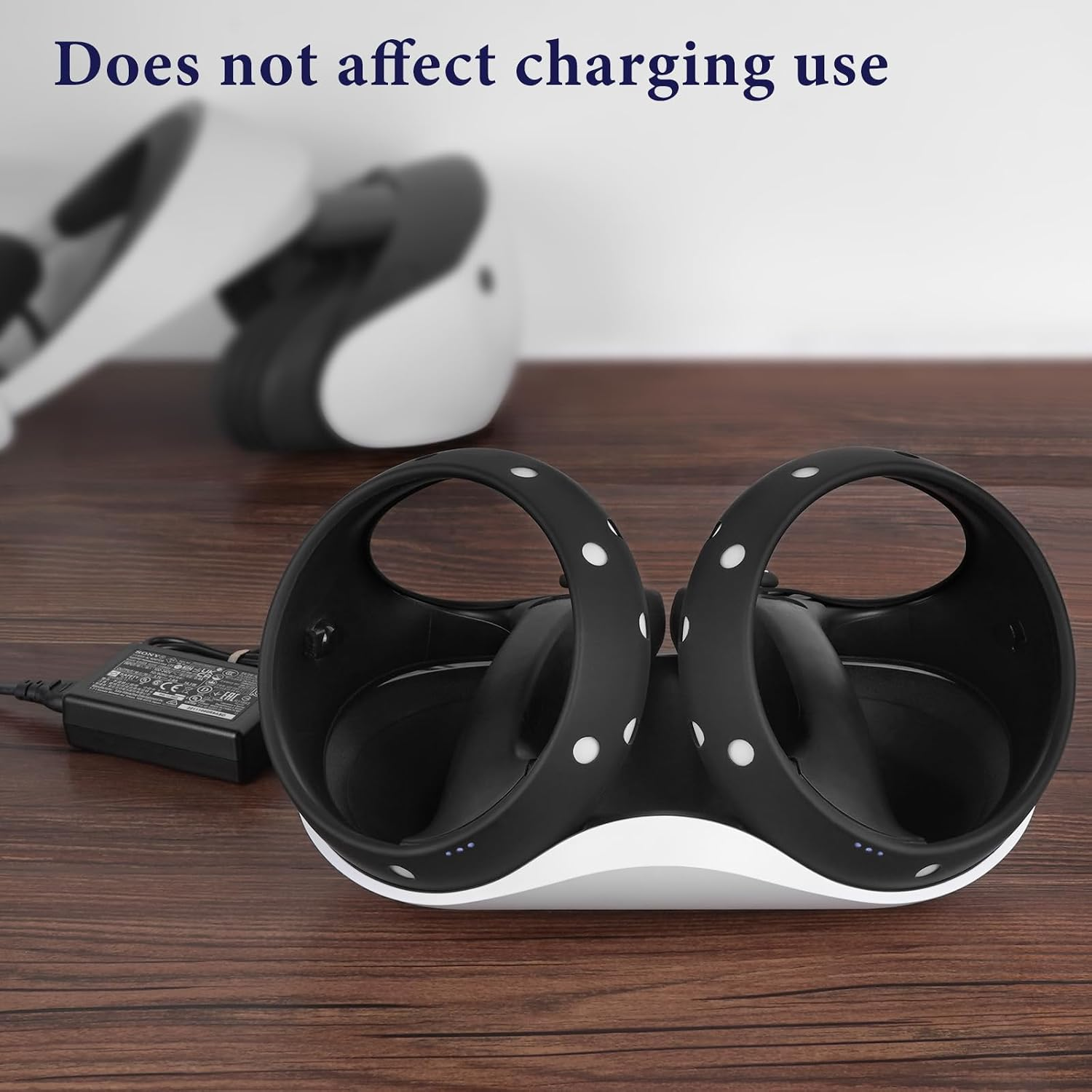 Zwei schwarze VR-Controller in einer weißen Ladestation auf einem Holztisch, dahinter VR-Headsets unscharf.