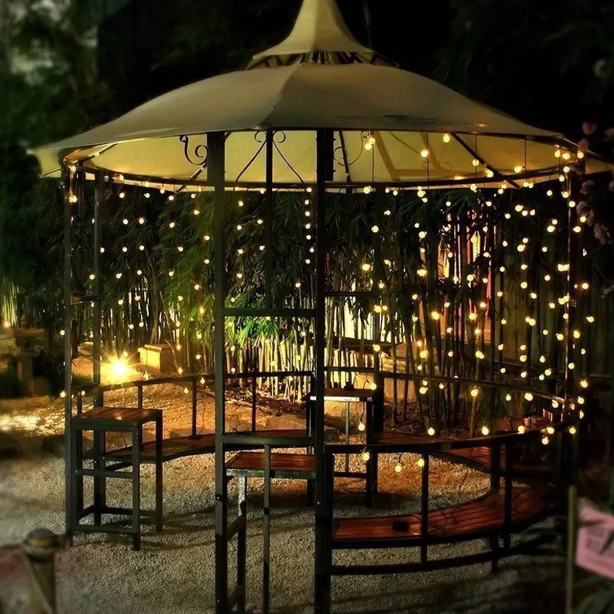 Ein beleuchteter Pavillon bei Nacht mit Lichterketten, Holzbänken und Bambusbäumen im Hintergrund.