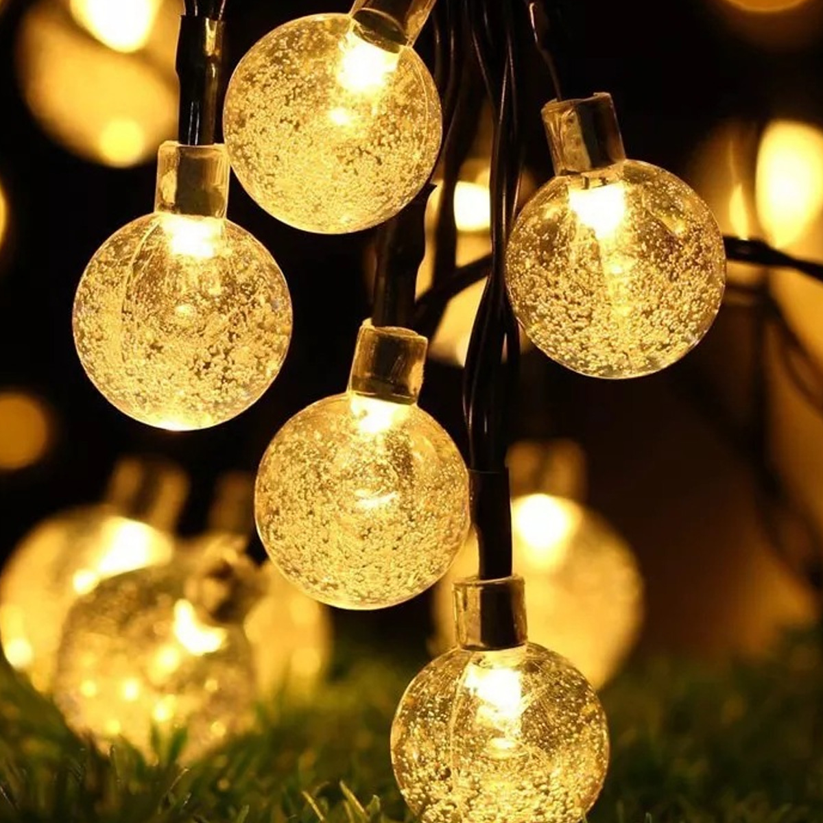 Leuchtende Kristallkugeln mit goldenem Licht hängen an dunklen Kabeln über Gras.