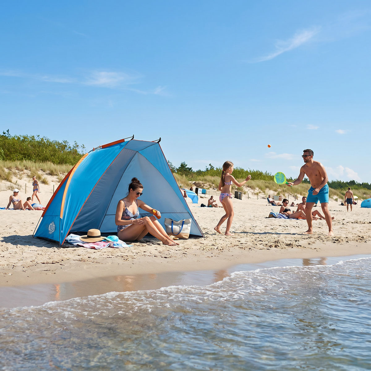 Kobieta siedzi w niebieskim namiocie plażowym, a rodzina gra w piłkę na plaży.