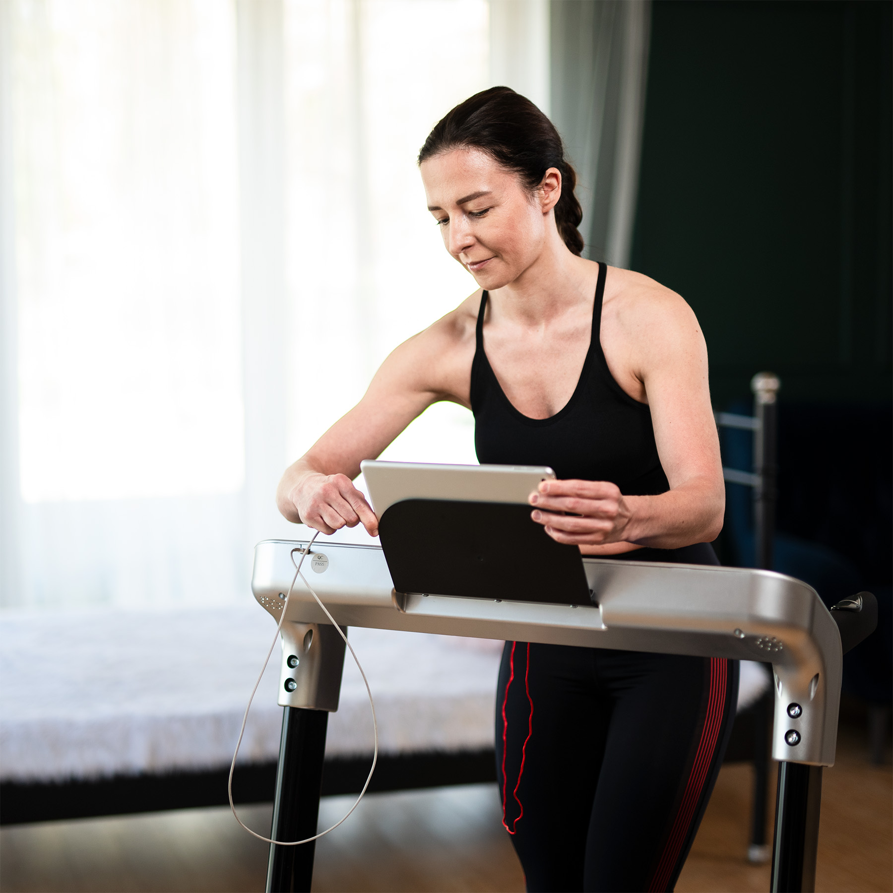 Kobieta w czarnym topie sportowym stoi na bieżni i obsługuje tablet.