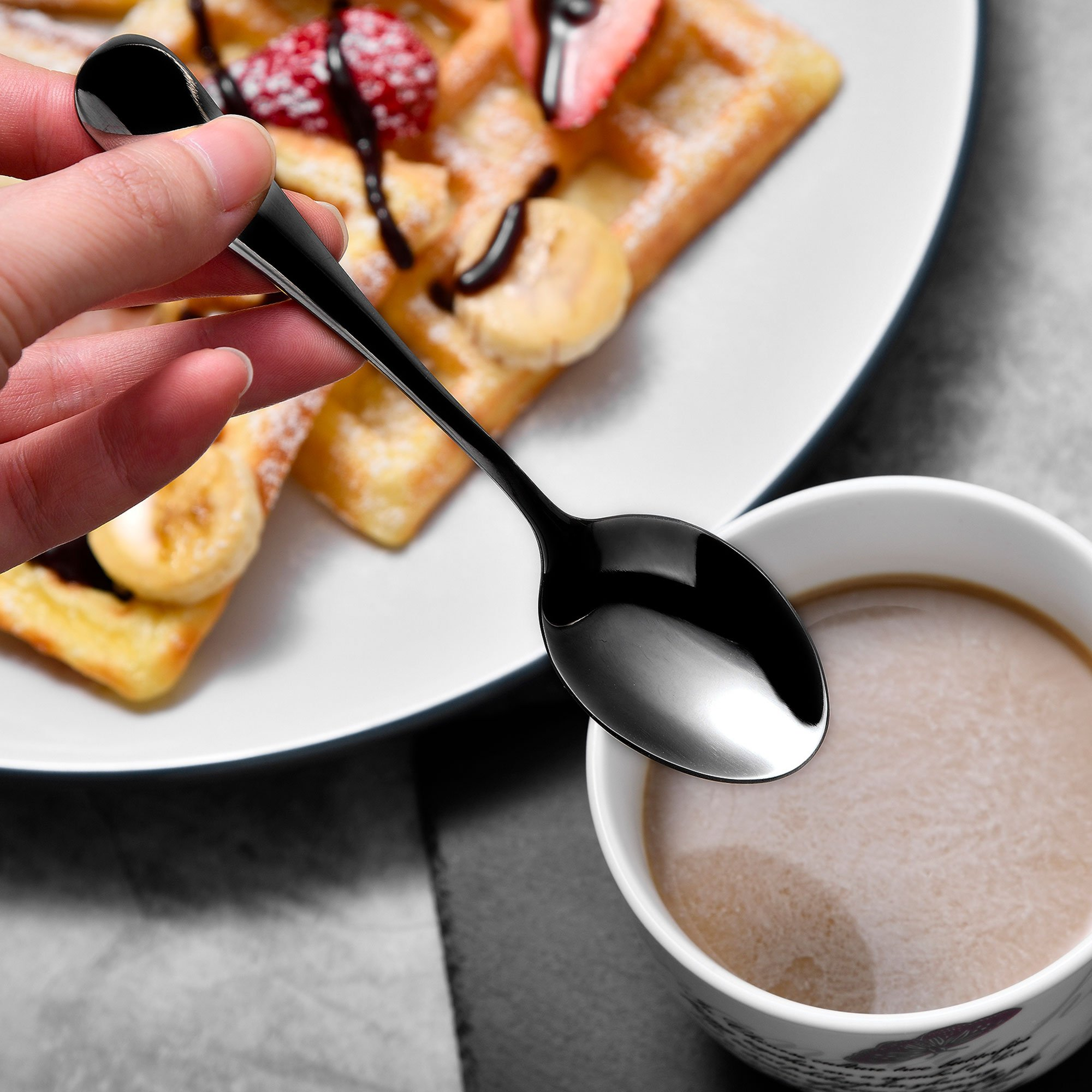 Una mano sostiene una cuchara negra sobre una taza de café, con gofres con fruta y chocolate al fondo.