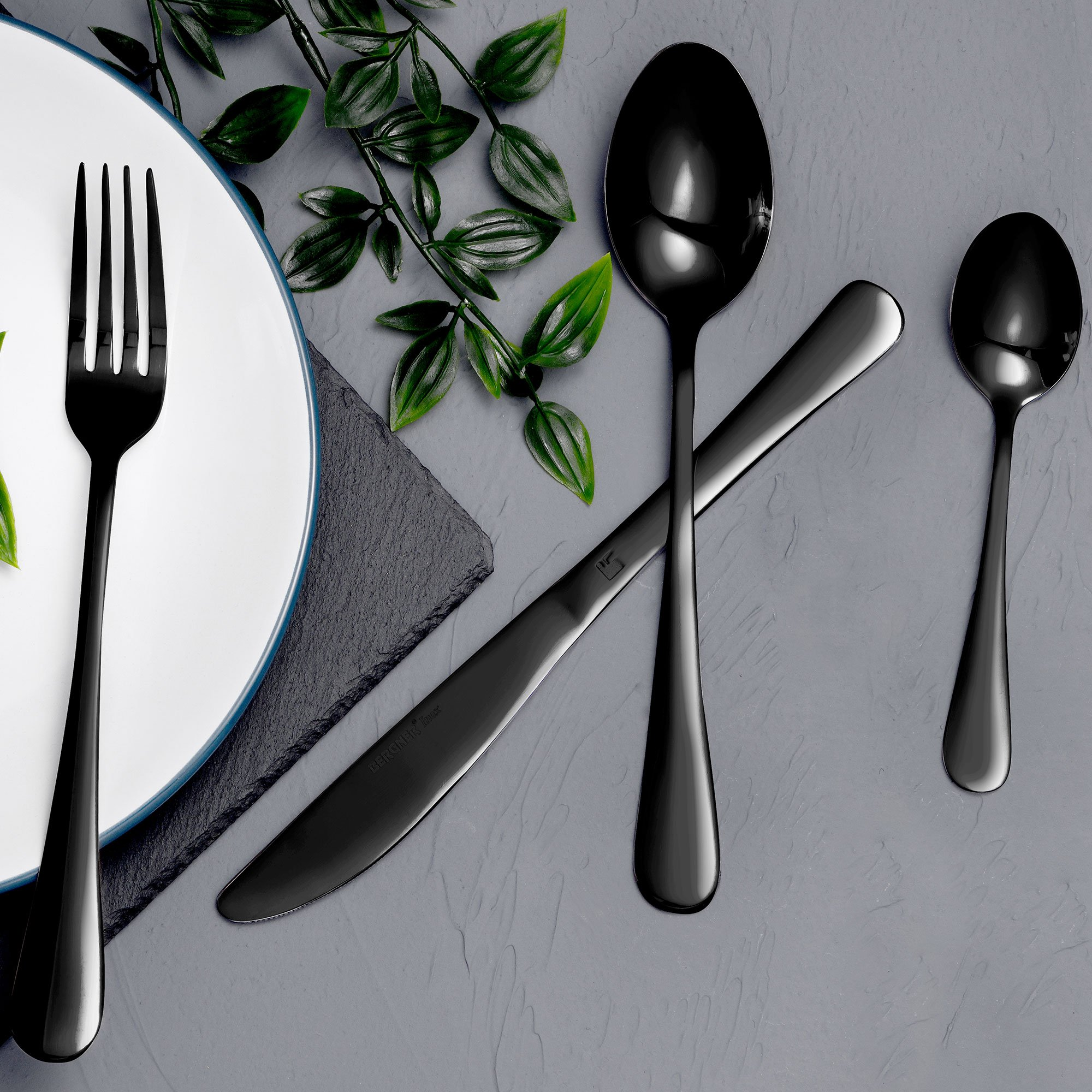 Juego de cubiertos negros con tenedor, cuchillo y cucharas sobre fondo gris.