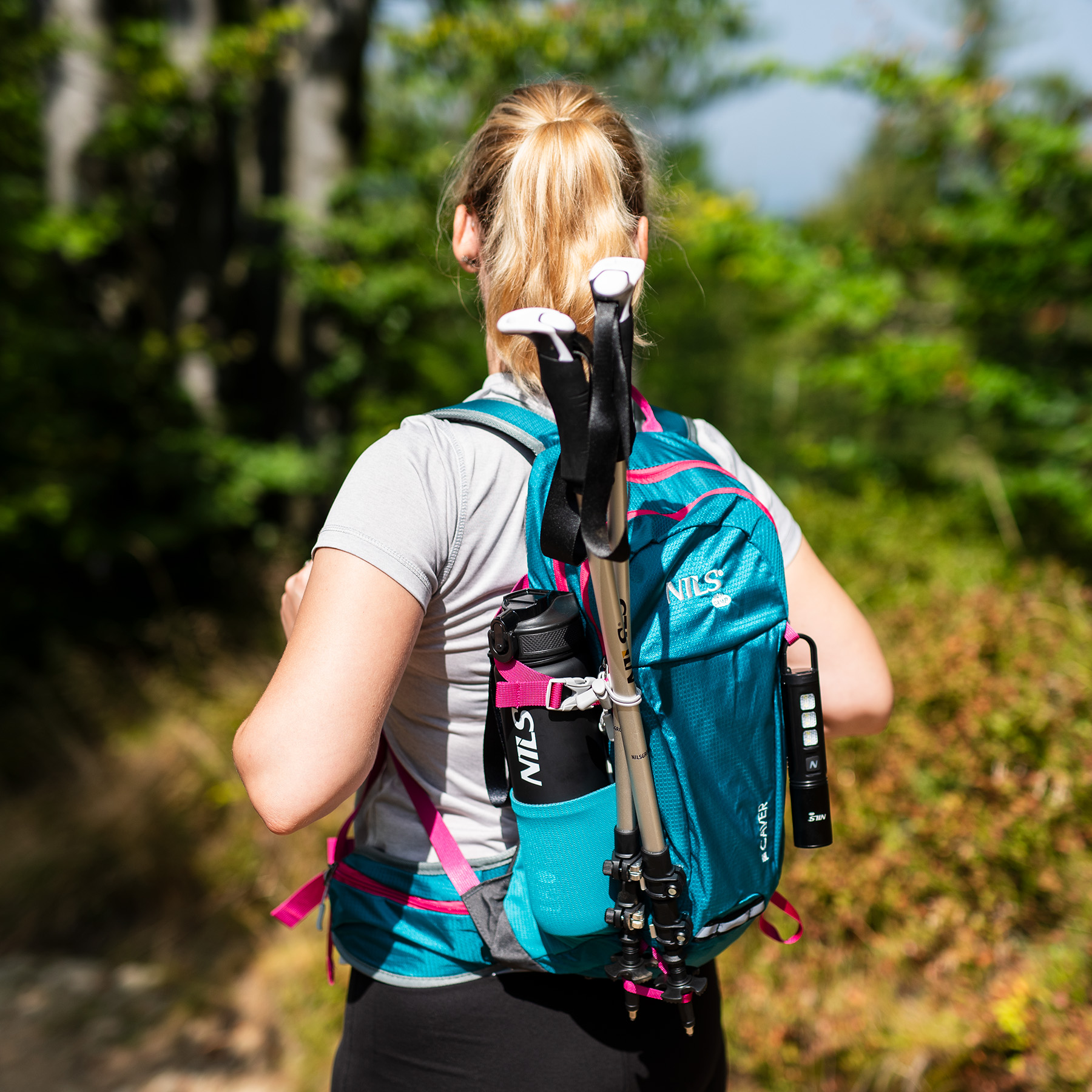 Blond kobieta od tyłu w niebieskim plecaku z kijkami trekkingowymi i bidonem, otoczona drzewami.