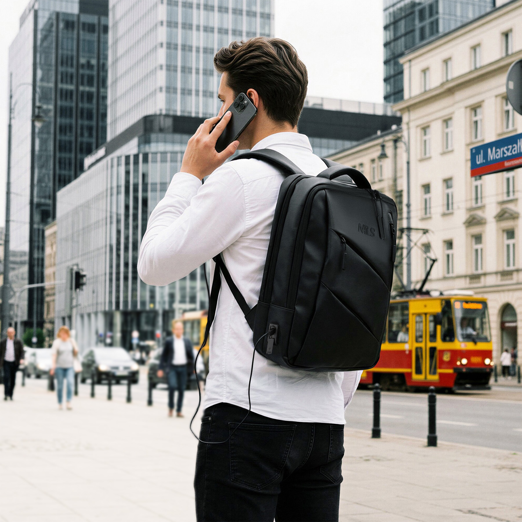 Mężczyzna z czarnym plecakiem i w białej koszuli stoi na ulicy miasta, rozmawiając przez telefon.