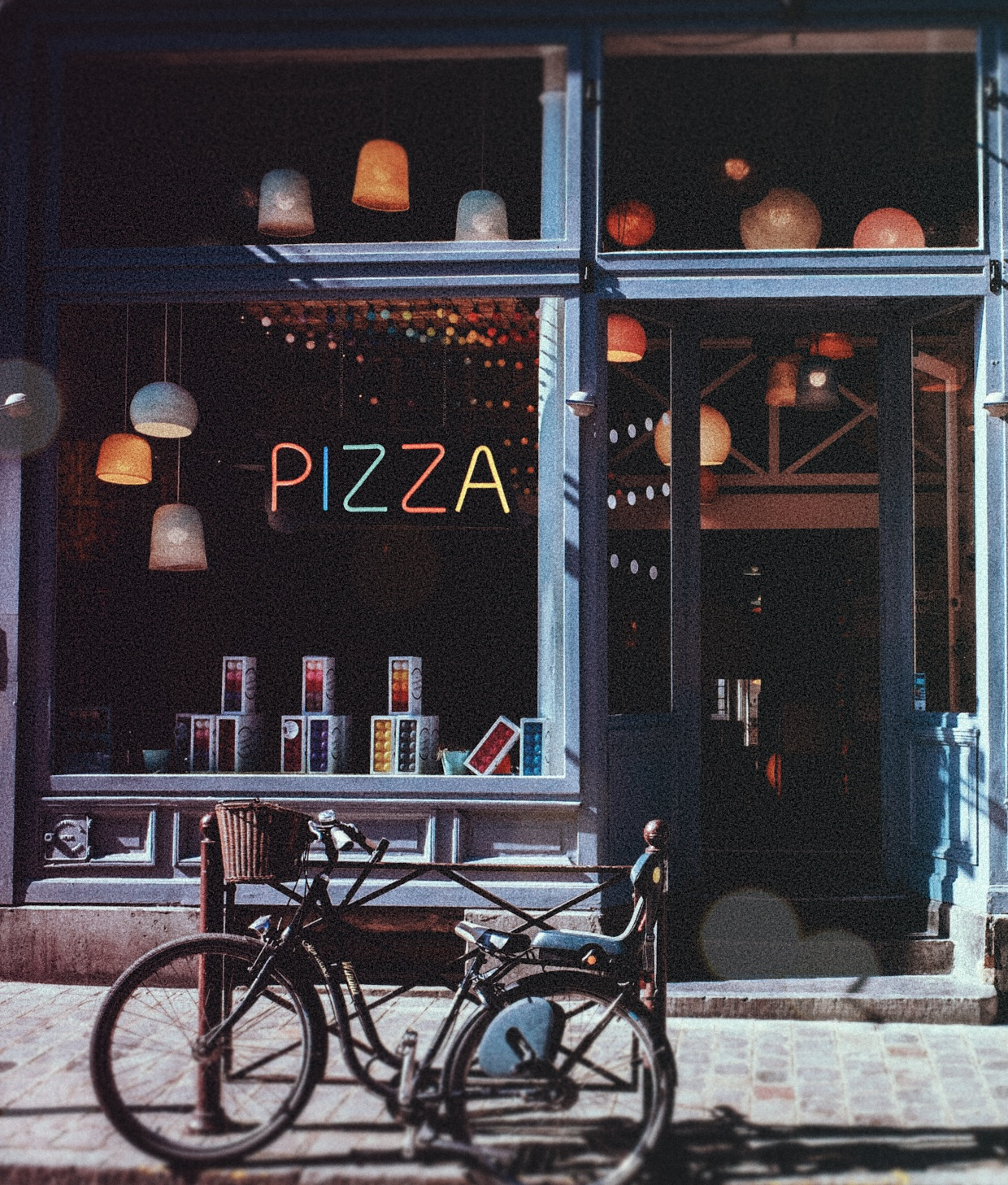 Eine Pizzeria mit bunten Neonschildern und Lampen, eine Fahrrad steht draußen auf dem Bürgersteig.