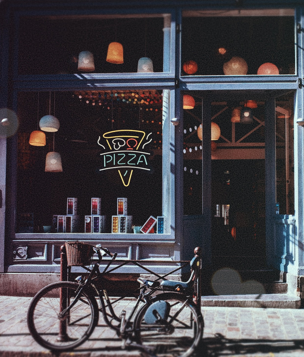 Ein Fahrrad steht vor einer Pizzeria mit Neonschild, leuchtenden Innenlampen und Schachteln im Schaufenster.