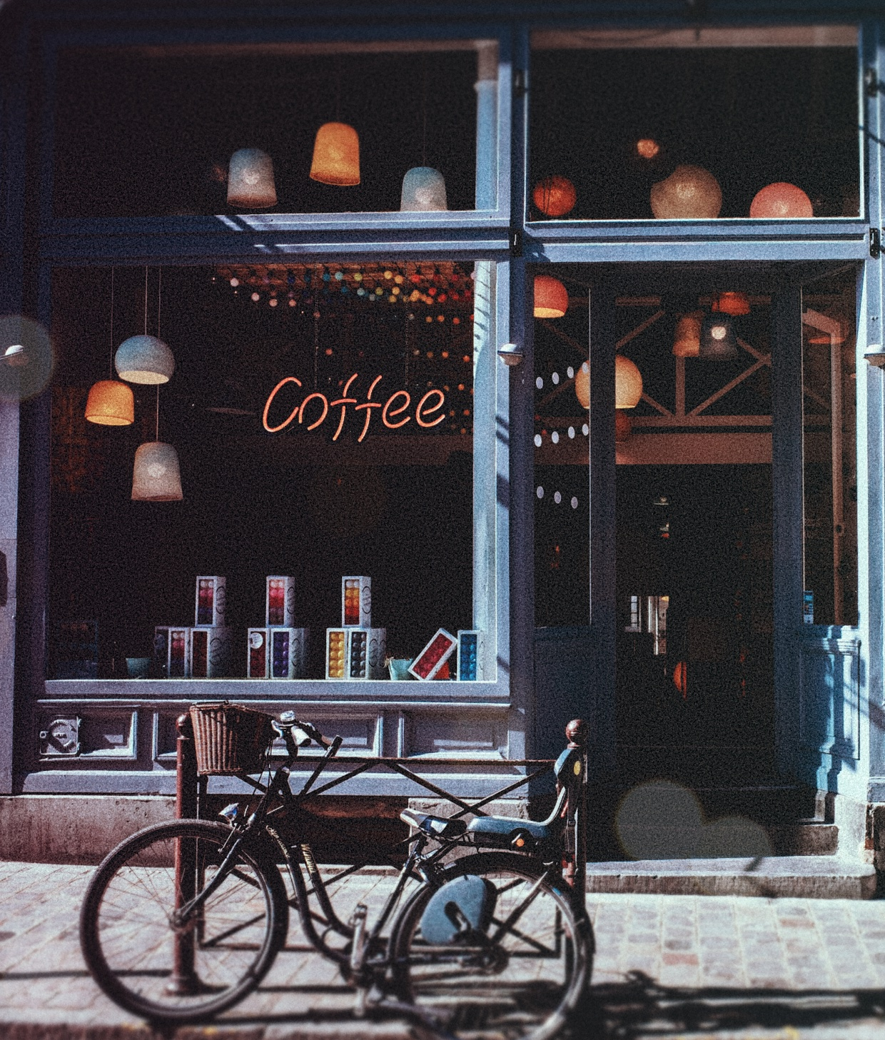 Gemütliches Café mit beleuchtetem 'Coffee'-Schild, hängenden Lampen, Produktboxen im Fenster und einem Fahrrad davor.