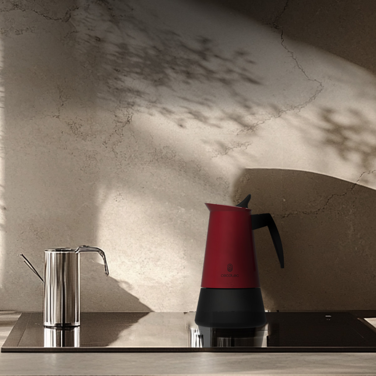 Una cafetera roja y un recipiente de metal plateado están sobre una placa oscura frente a una pared texturizada.