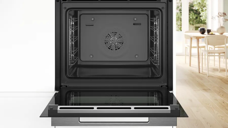 Un forno nero aperto e vuoto con griglie laterali, con una cucina luminosa sullo sfondo.