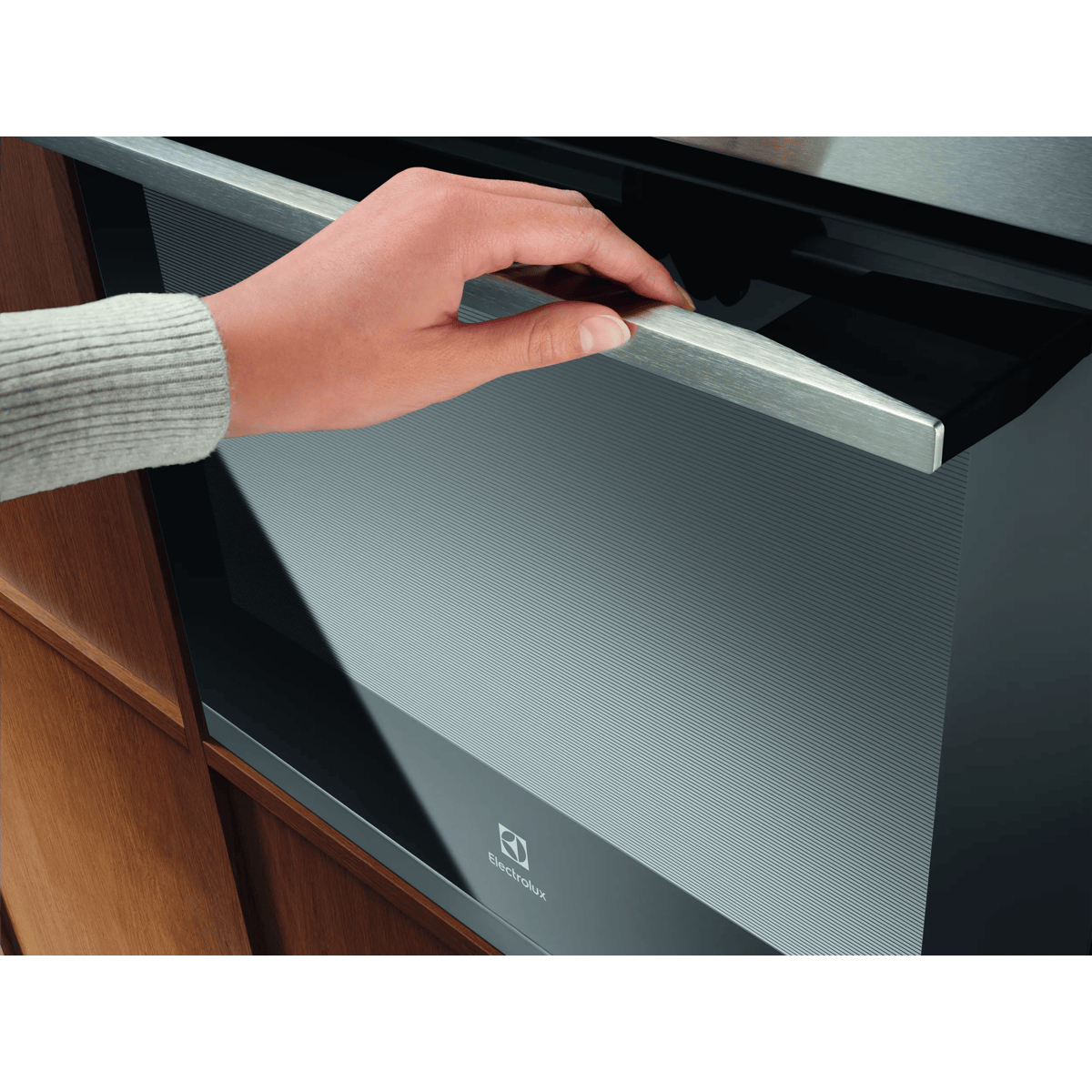 Una mano apre la maniglia in acciaio inossidabile di un forno Electrolux nero in una cucina in legno.