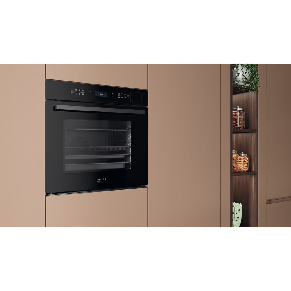 Un forno da incasso Hotpoint nero in una cucina beige accanto a uno scaffale con barattoli e piante.