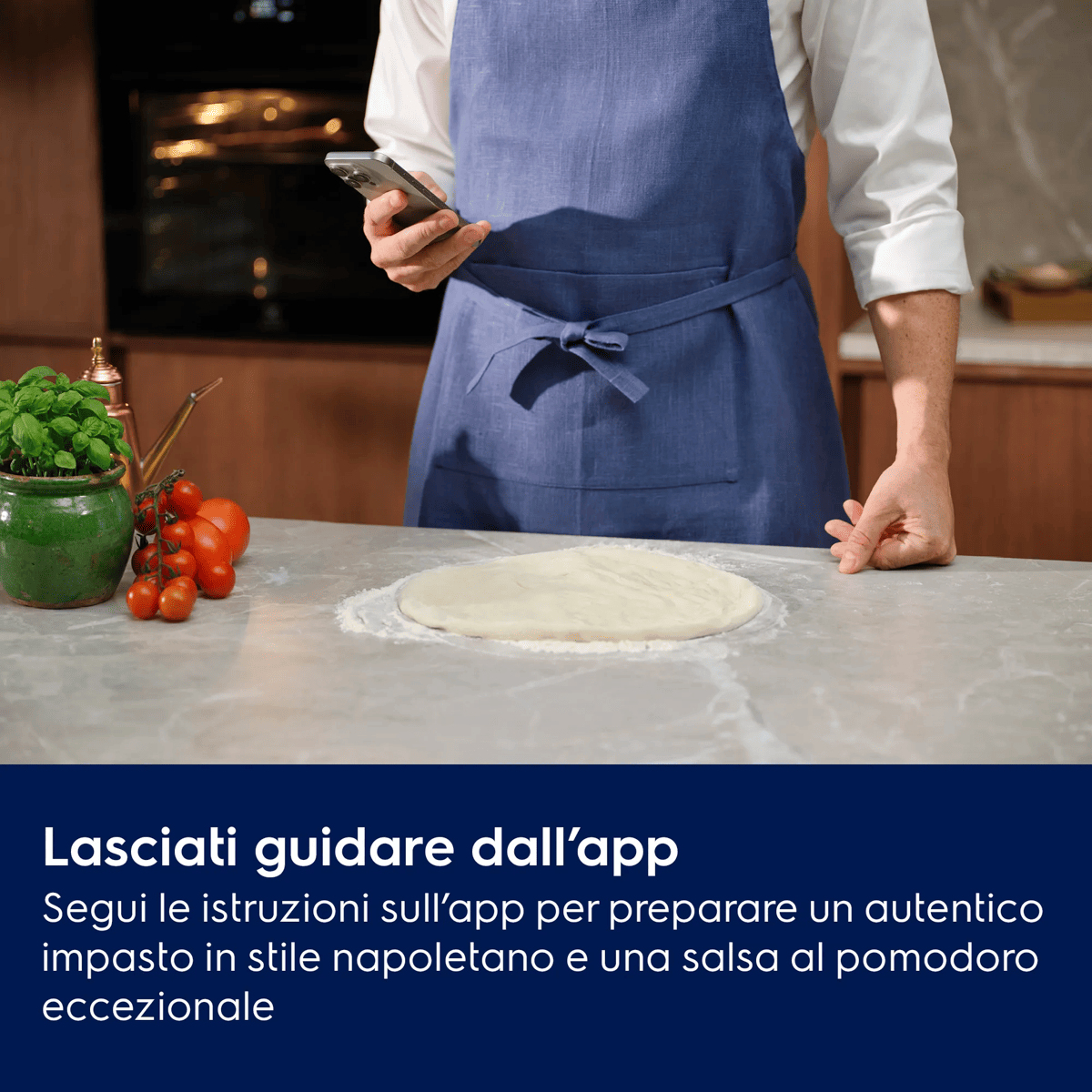Una persona con grembiule blu tiene uno smartphone vicino all'impasto su un bancone con basilico e pomodori.