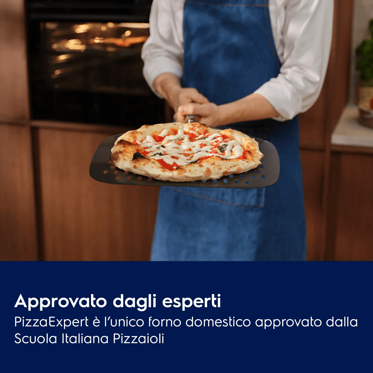 Una persona con grembiule blu tiene una pizza fresca su una pala in una cucina.