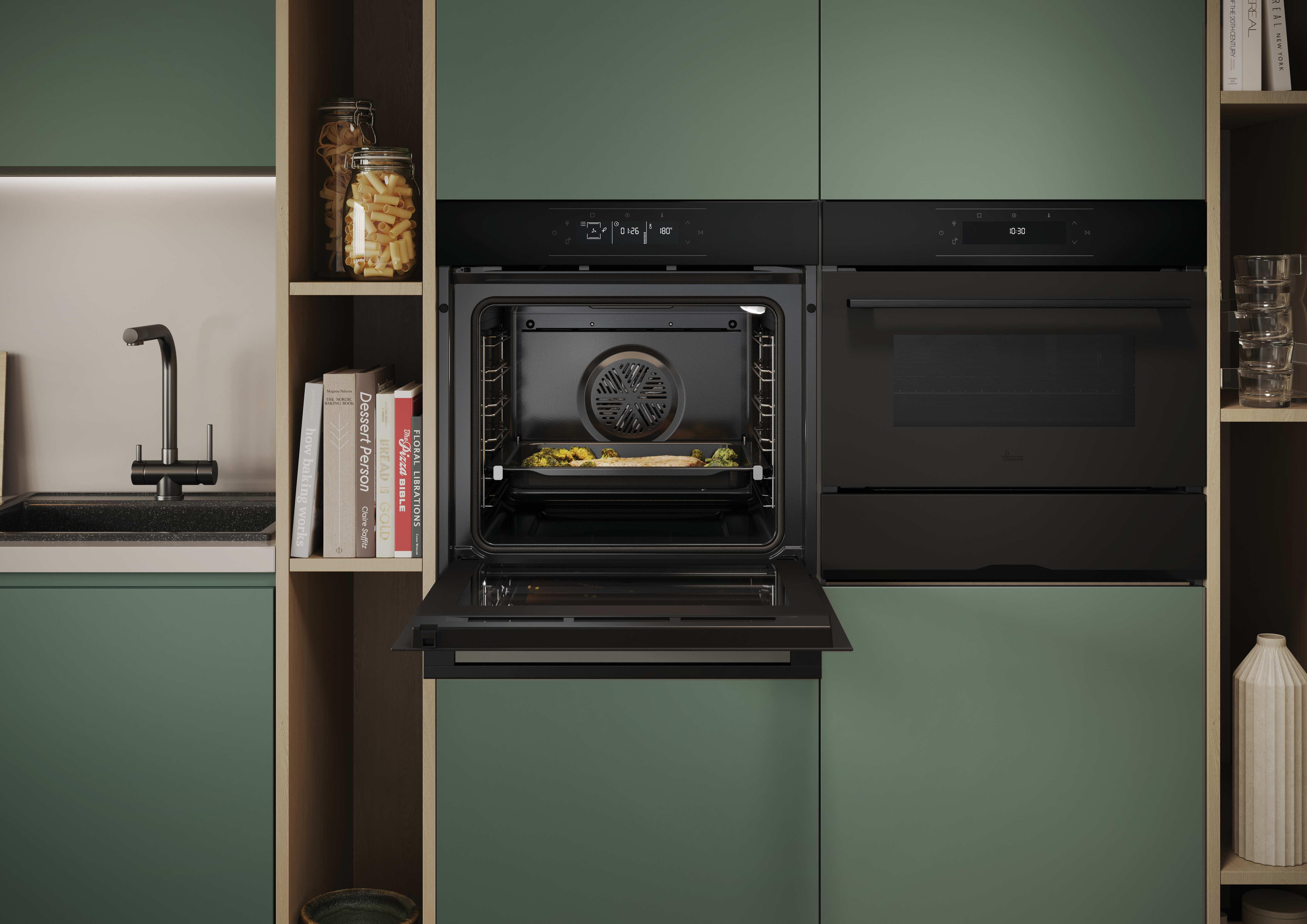 Una cucina moderna con mobili verdi, forni neri da incasso e un forno aperto che cuoce del cibo.