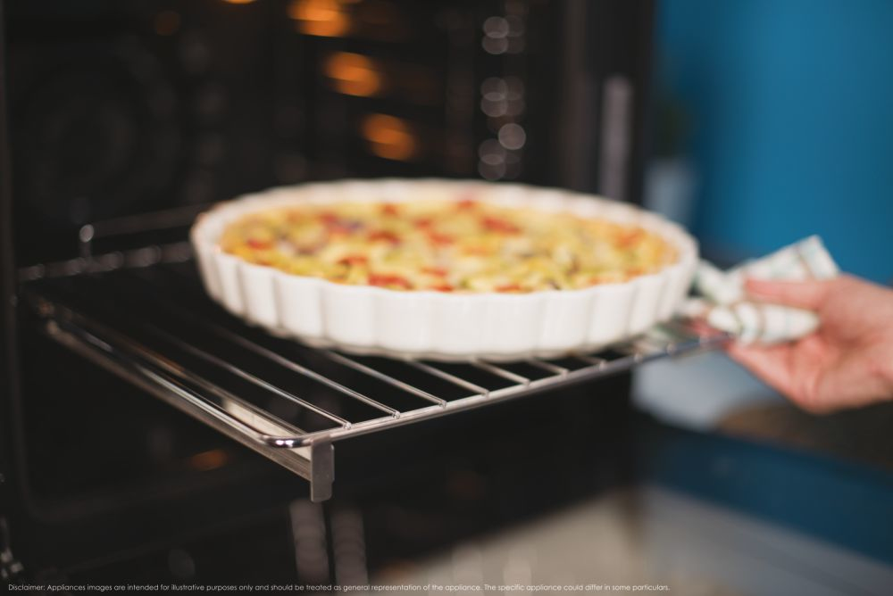 Una mano estrae dal forno una griglia con una quiche cotta in un piatto bianco.