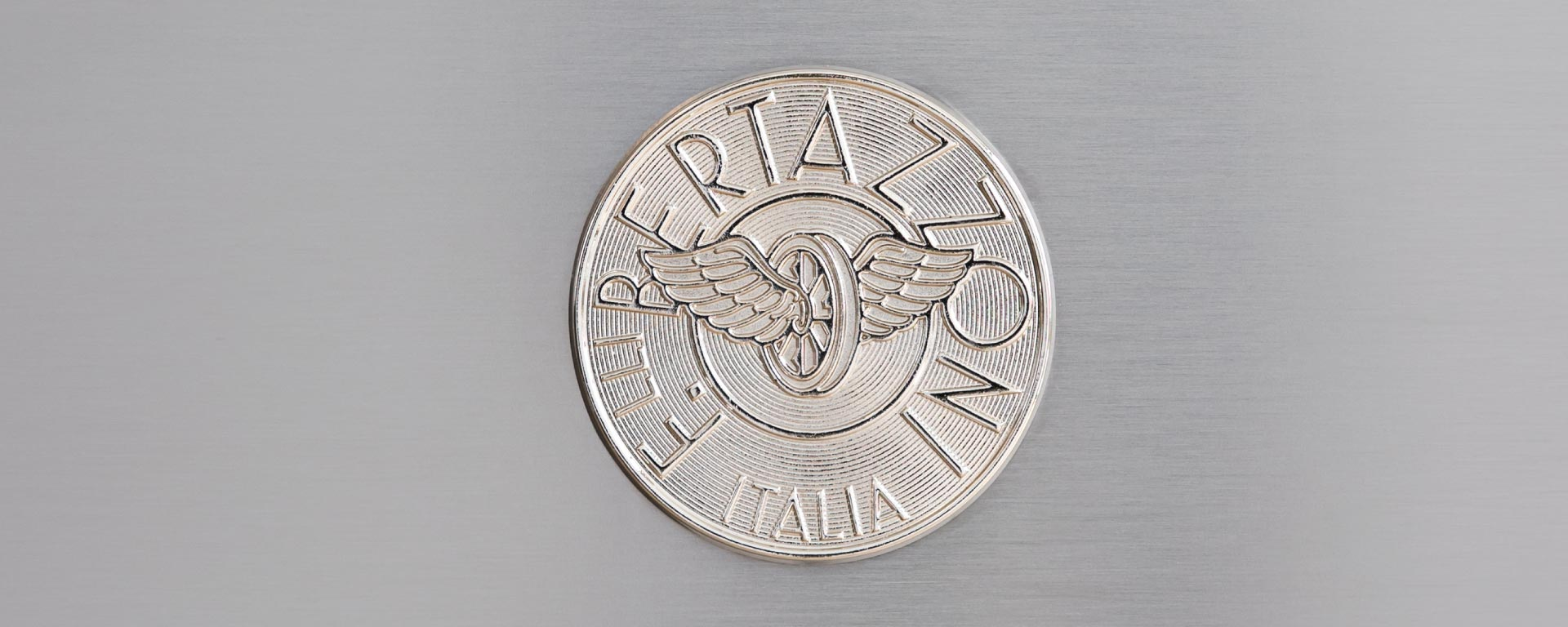 Logo argentato Bertazzoni con una ruota alata su metallo spazzolato.