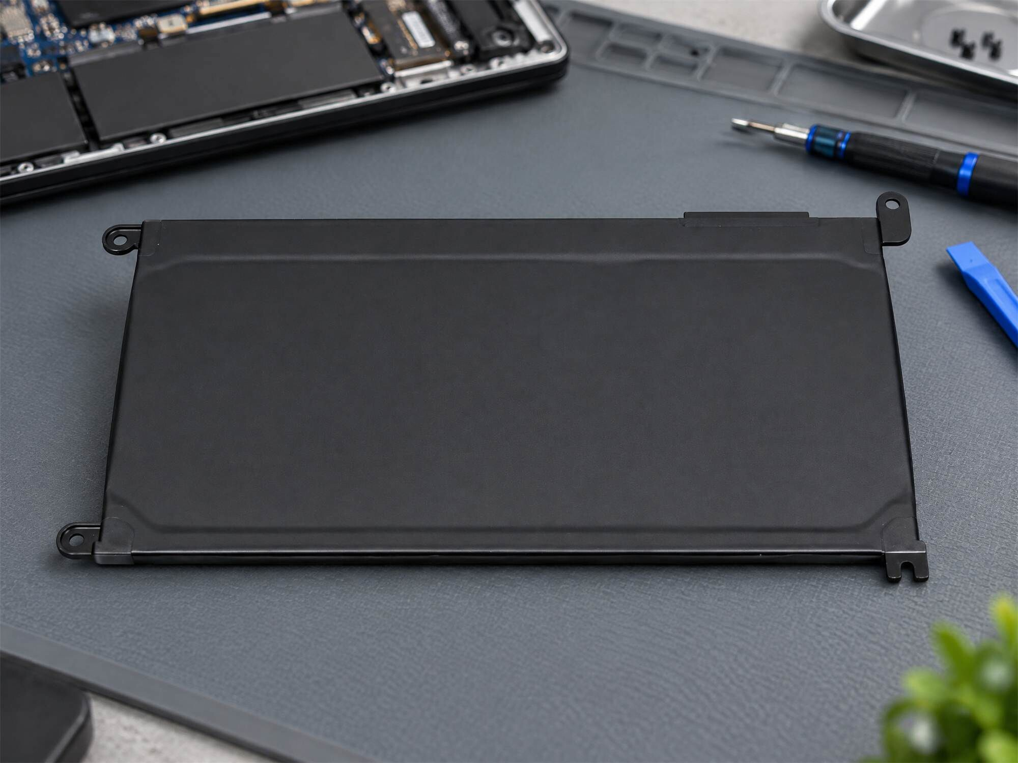 Ein schwarzer Laptop-Akku liegt auf einer grauen Reparaturmatte mit Werkzeugen und Laptop-Teilen im Hintergrund.