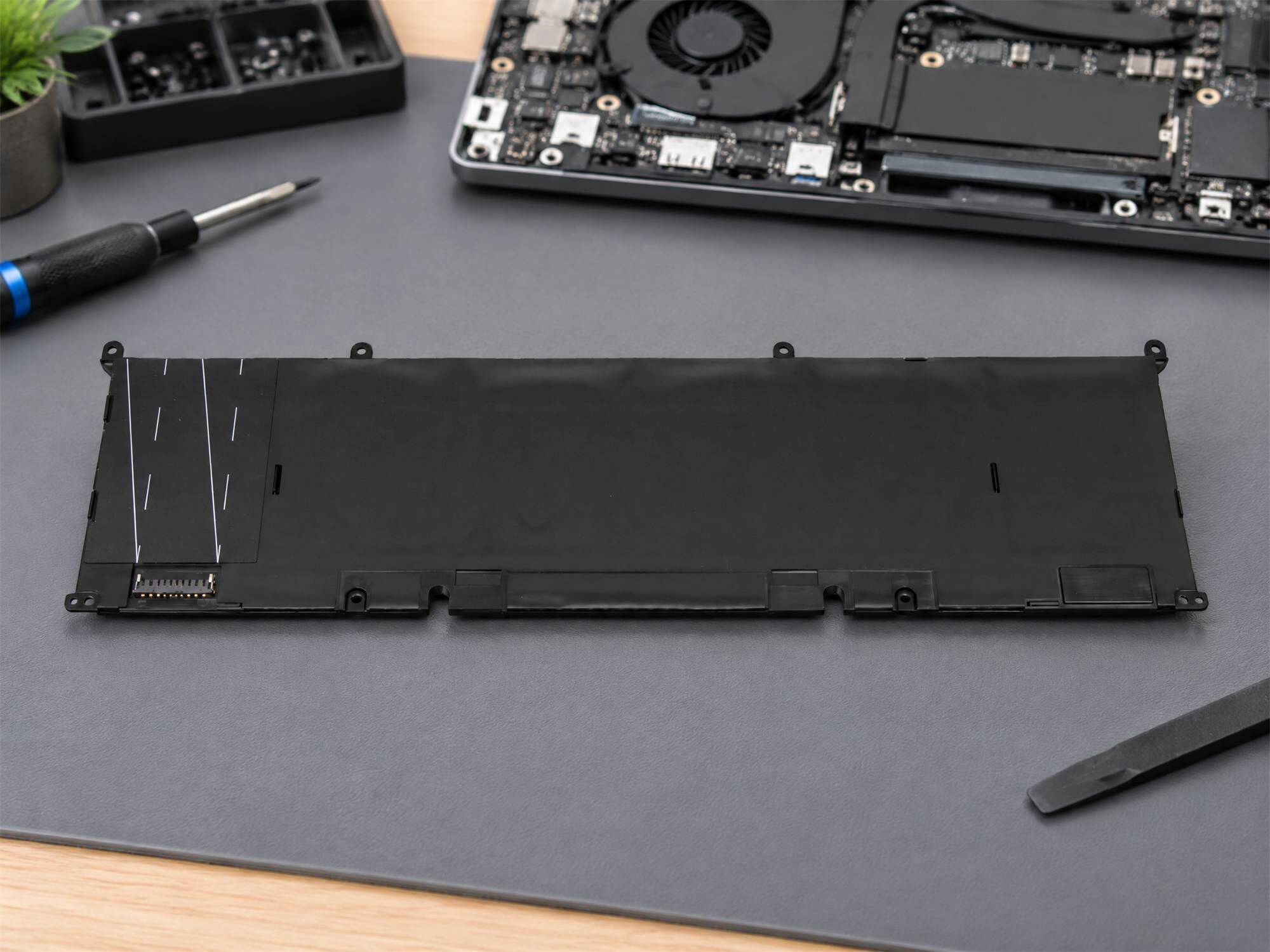 Flacher schwarzer Laptop-Akku auf einer grauen Matte, umgeben von Werkzeugen und einem geöffneten Laptop.